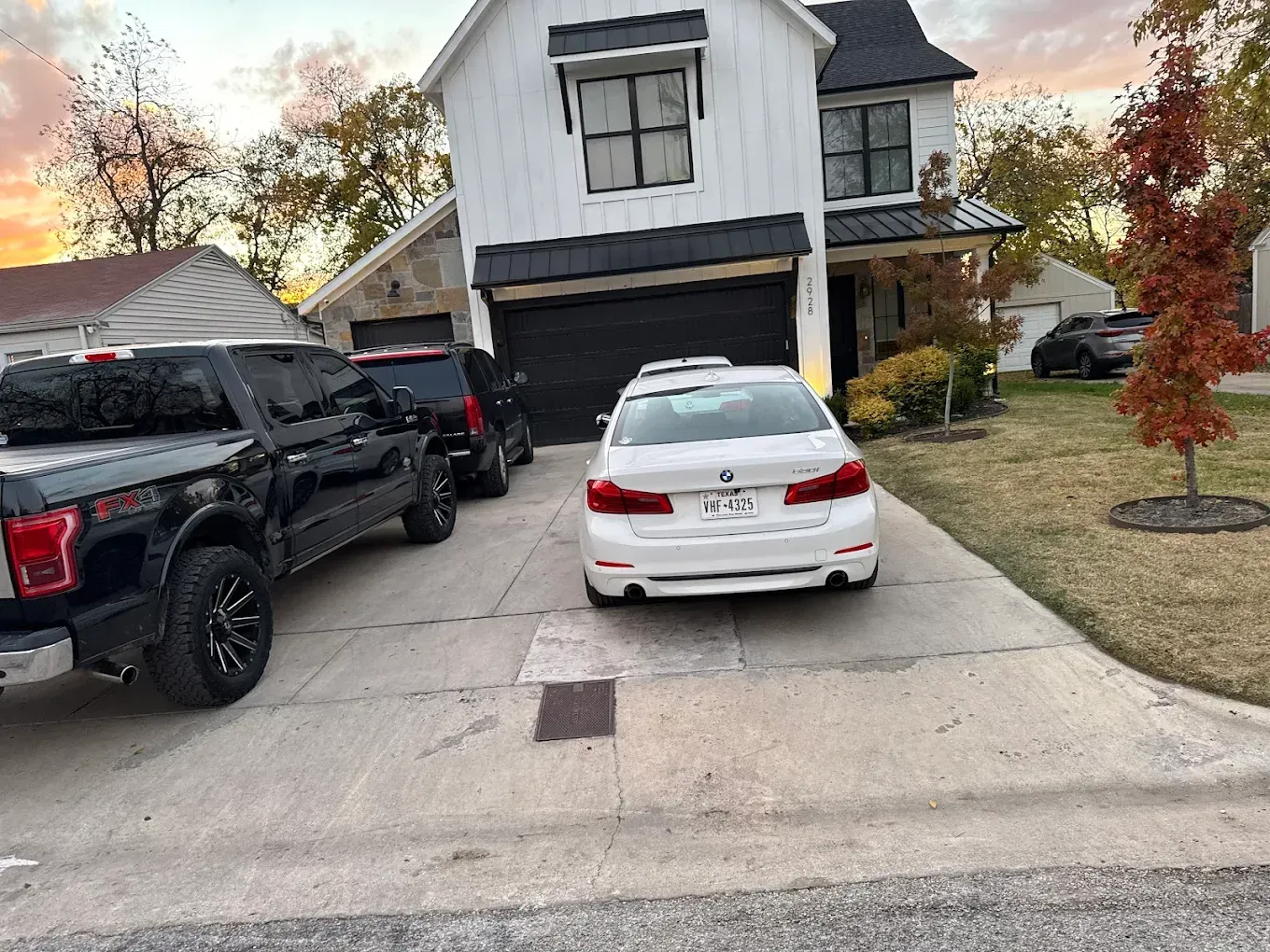 White car parked on driveway in front of two-story white house with black garage door, and a black truck parked next to it.