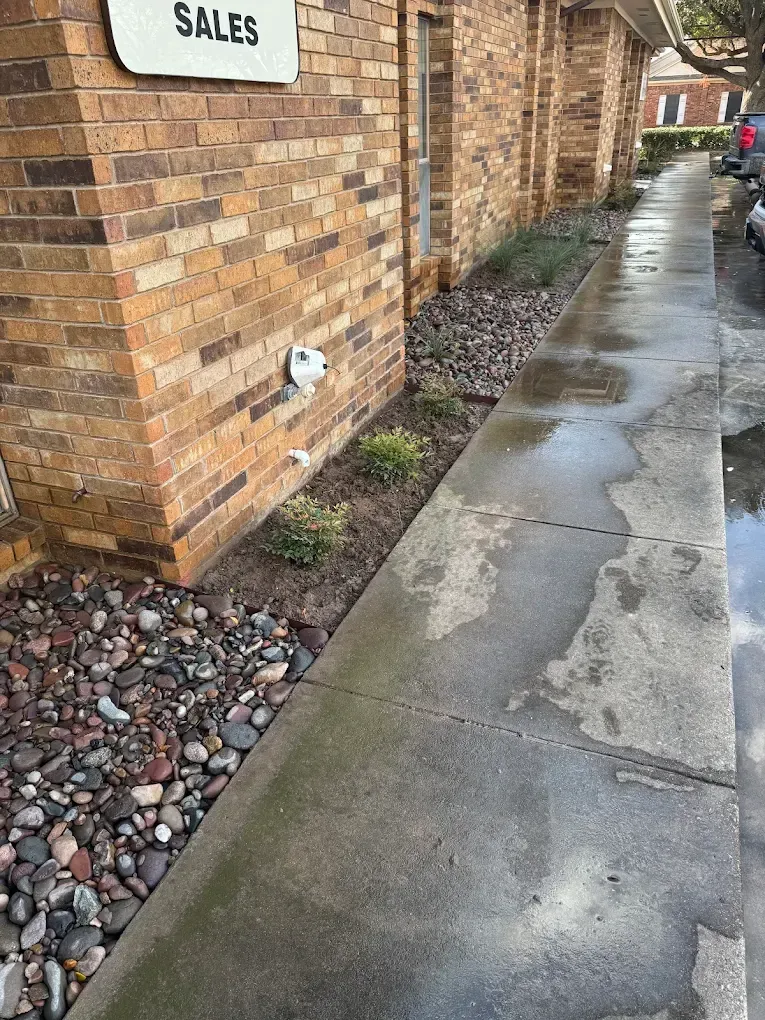 Brick building exterior with a narrow flower bed and wet sidewalk.