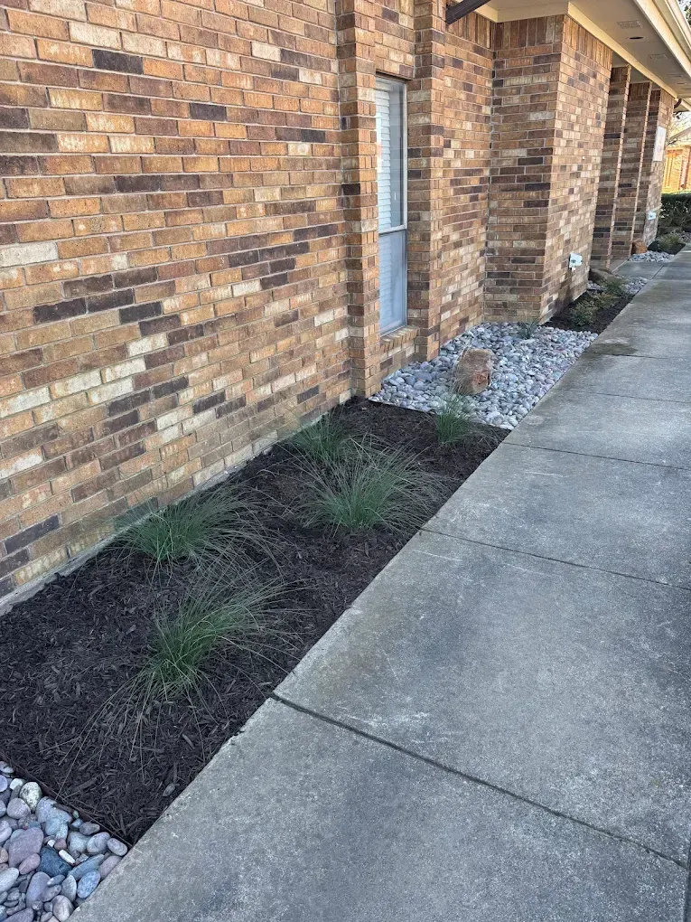 A brick building with a concrete walkway and a landscaped area with mulch, plants, and rocks.