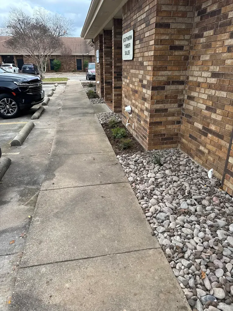 Sidewalk beside a brick building with parking spaces. Landscaping includes rocks and plants.