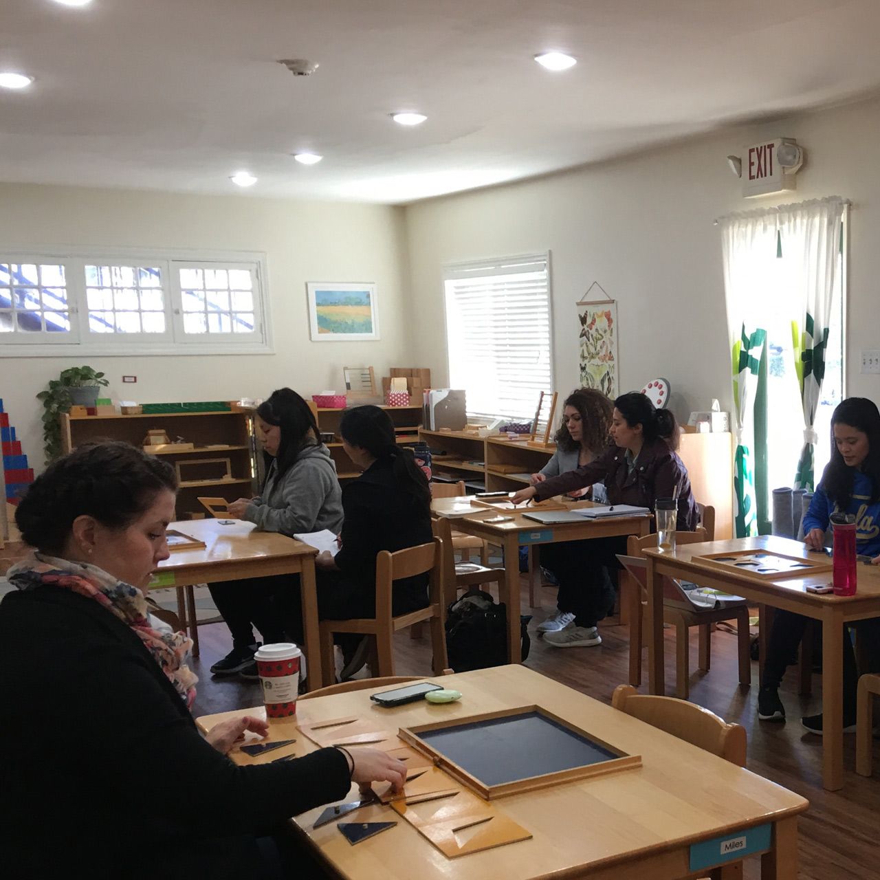 Adult students in an AMS training program practice using the Montessori geometric cabinet at wooden classroom tables.