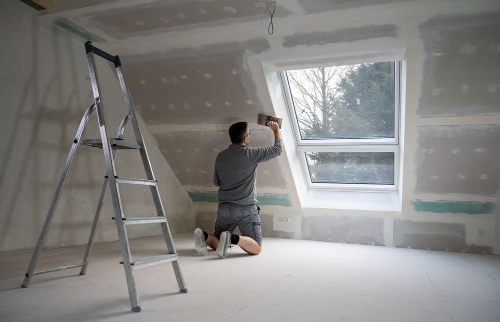 A Man is Kneeling Down in Front of a Window in a Room With a Ladder — Mark's Flooring & Handyman Services in Erina, NSW
