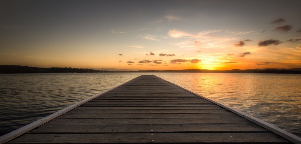 A Wooden Dock Leading Into a Body of Water at Sunset — Mark's Flooring & Handyman Services in Long Jetty, NSW