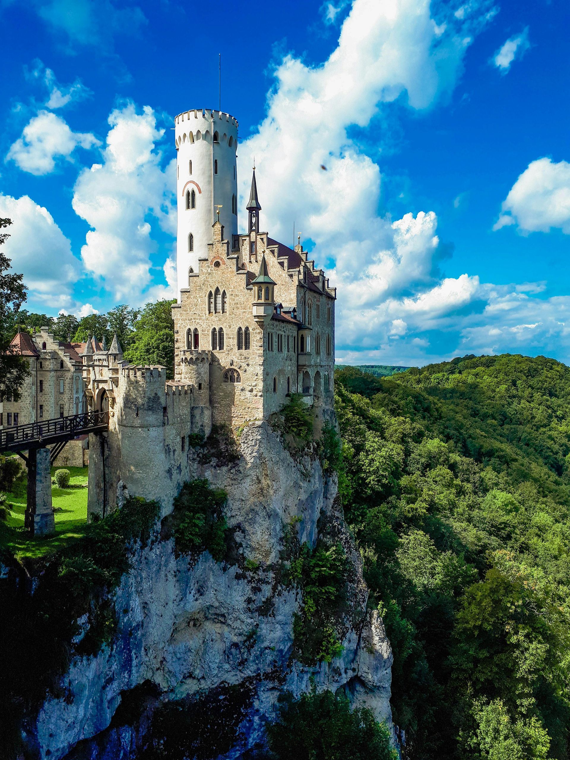 Castle Lichtenstein perched atop a cliff with a stone bridge and white tower, against a blue sky.