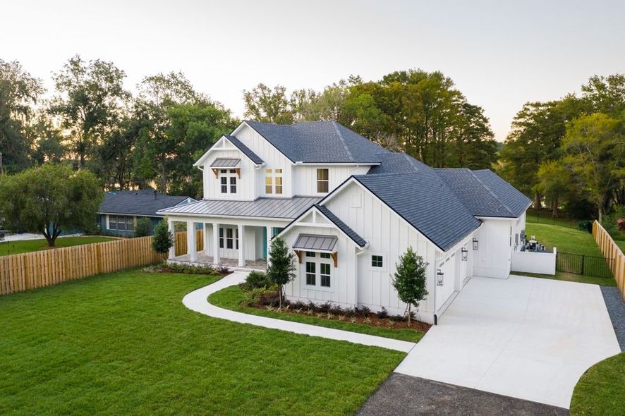 Two-story white farmhouse with black roof and gray driveway, on a green lawn with a wooden fence.