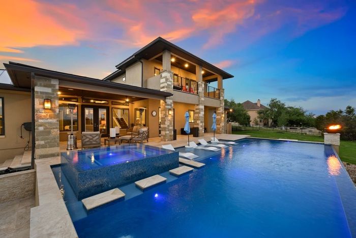 Luxury home with infinity pool at sunset. Beige exterior, blue water, colorful sky.