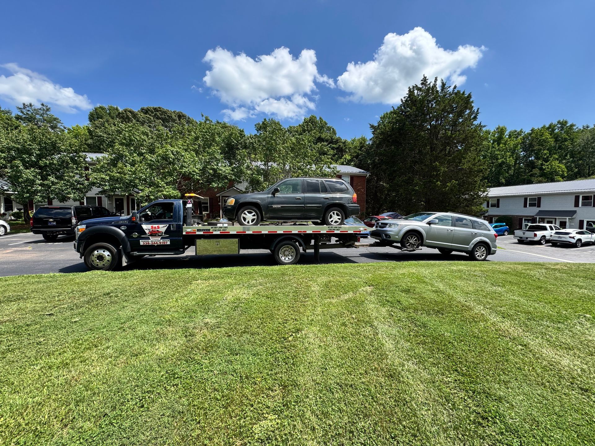 Tow truck carrying two SUVs on a grassy area, in front of a residential building on a sunny day.