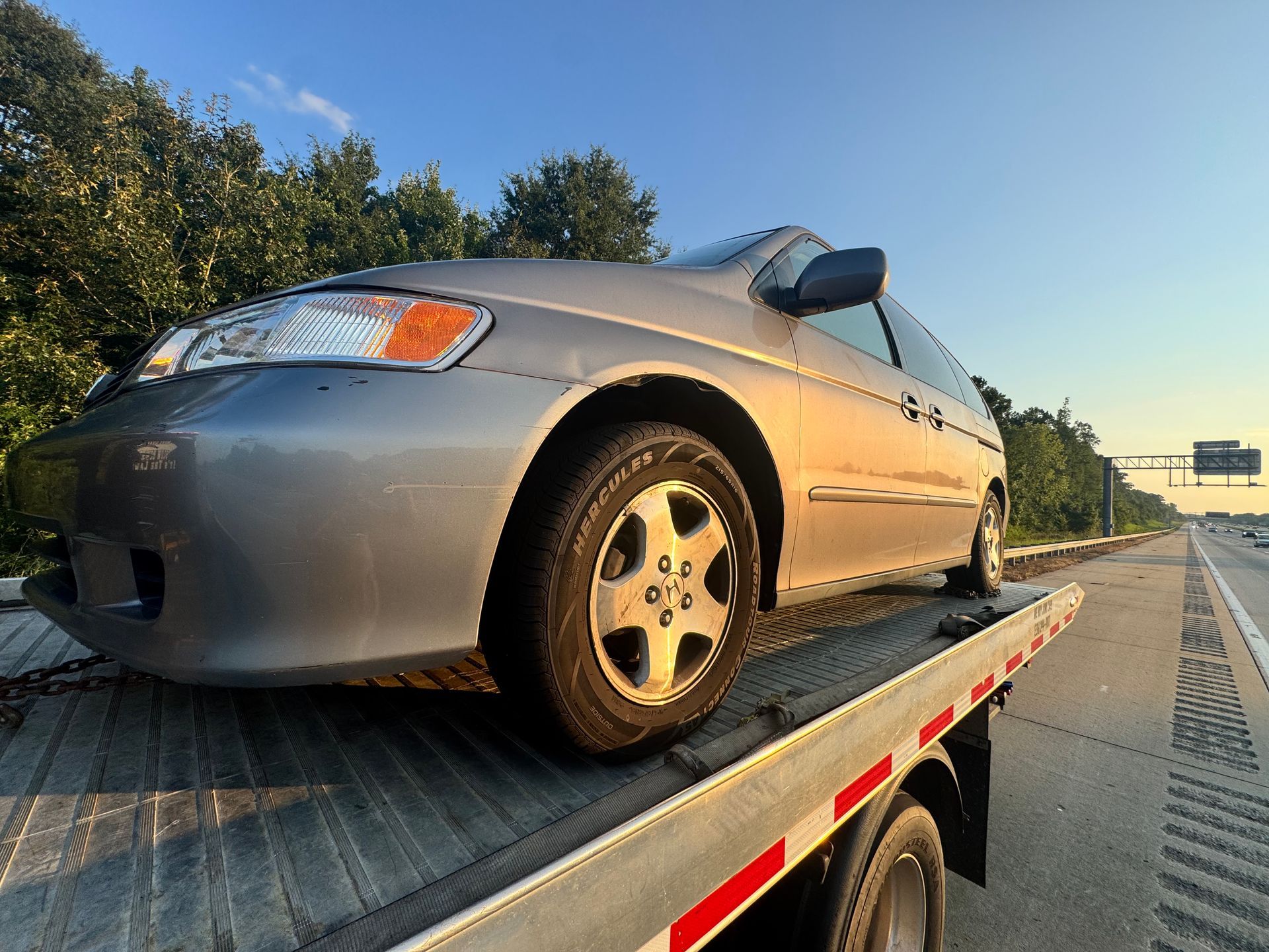 Silver car on a flatbed tow truck on a highway, with a blue sky background.
