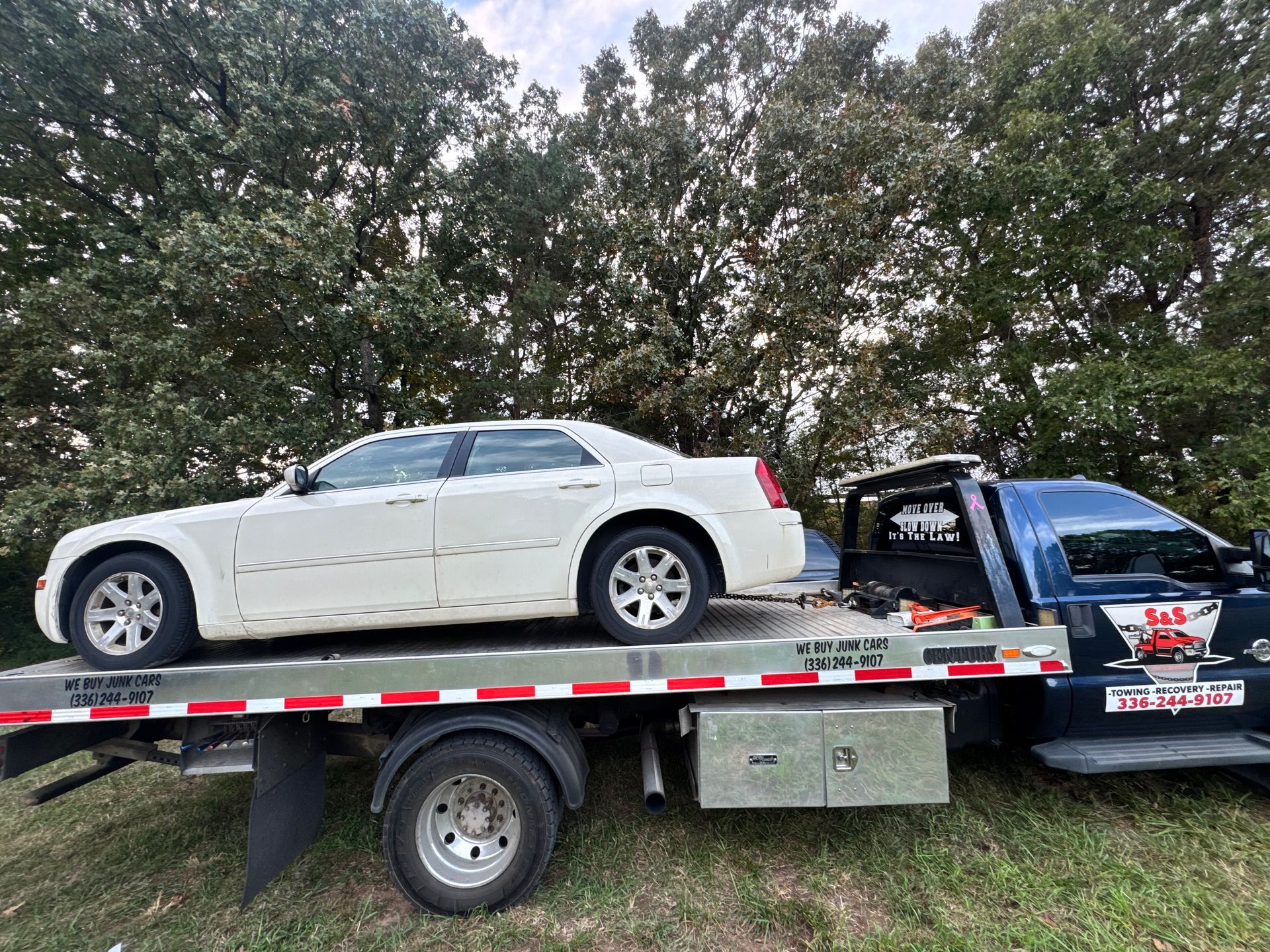 White car on a tow truck, parked near trees. Blue tow truck has BAS Towing logo on door.