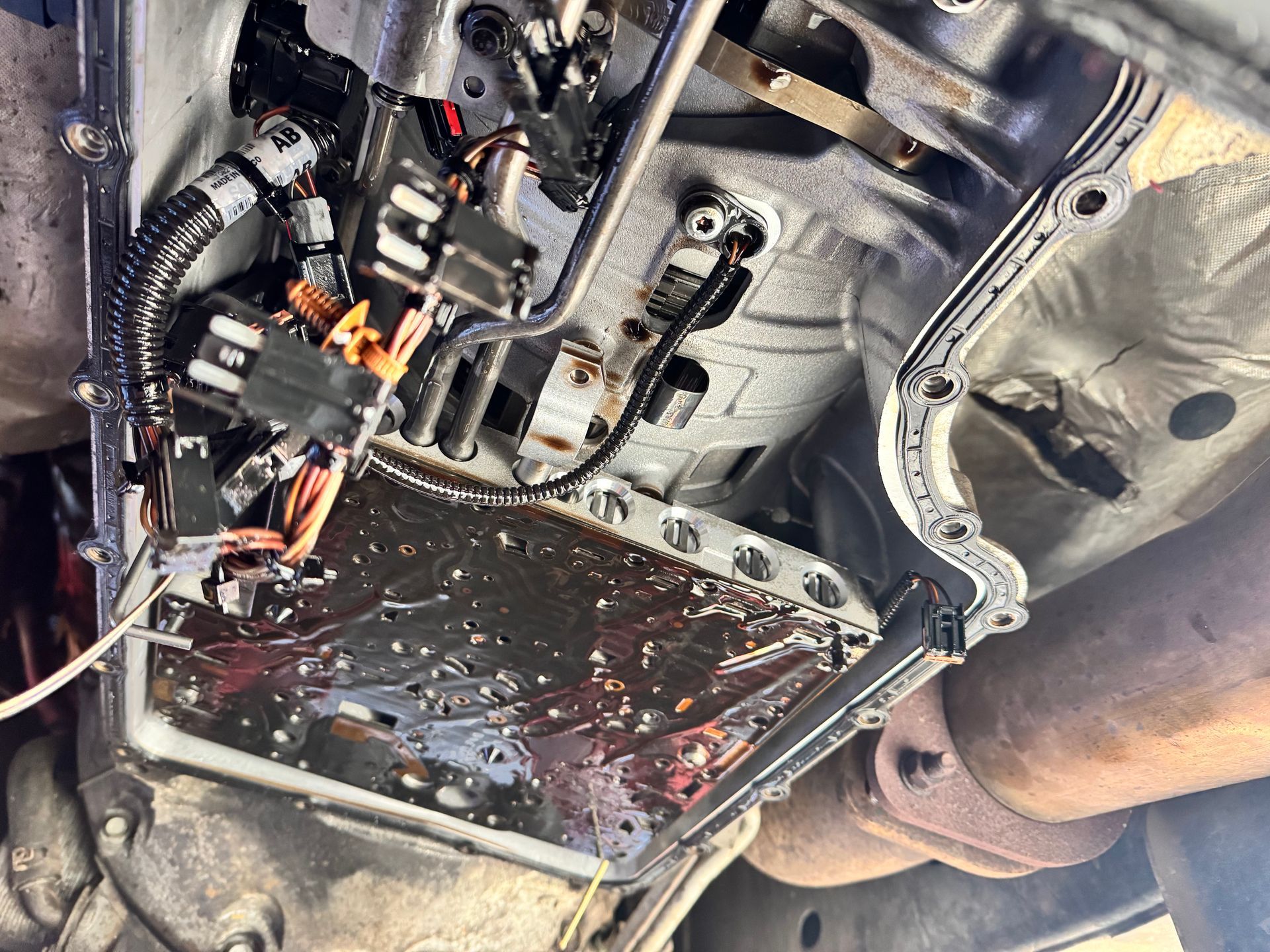 Underside of a vehicle, transmission pan removed, revealing dark fluid and components. A new gasket is in place.