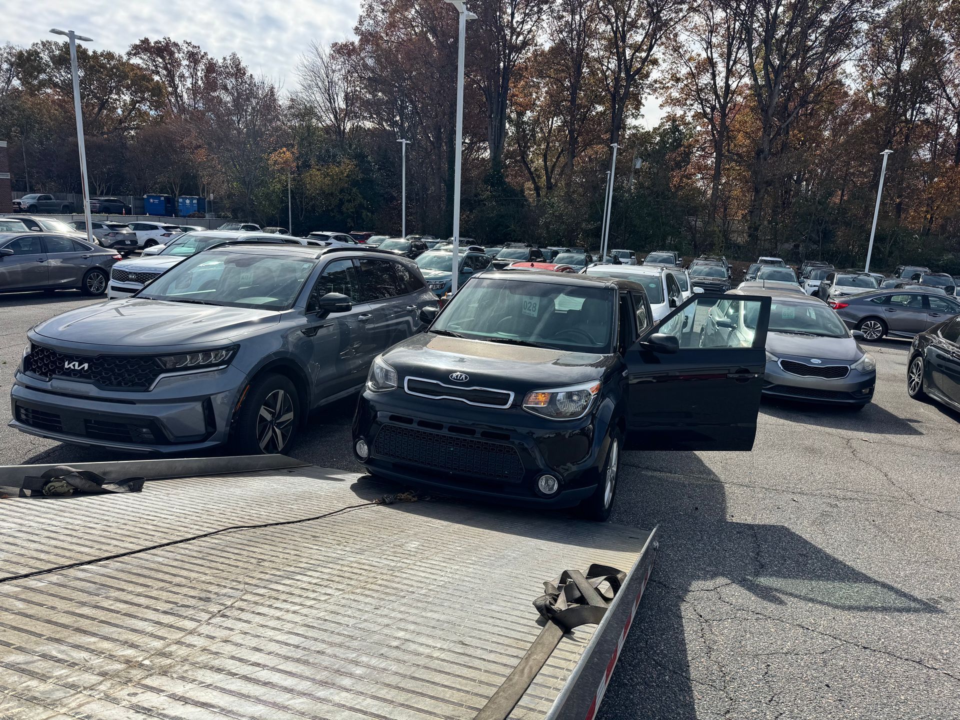 Two cars, one Kia Soul and a Kia SUV, on a car transport trailer in a car dealership lot.
