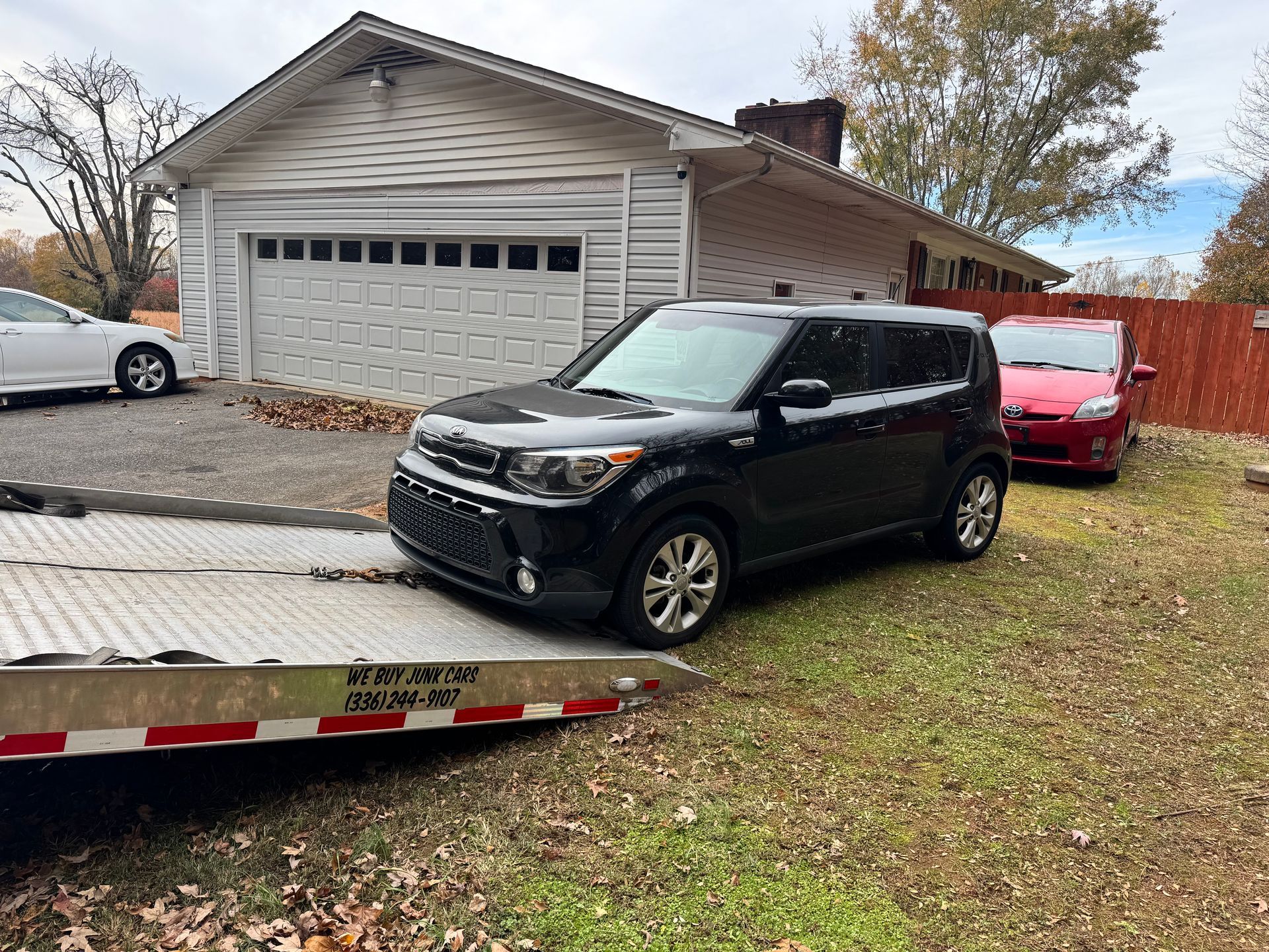Black Kia Soul on a tow truck ramp in front of a white garage and red car.