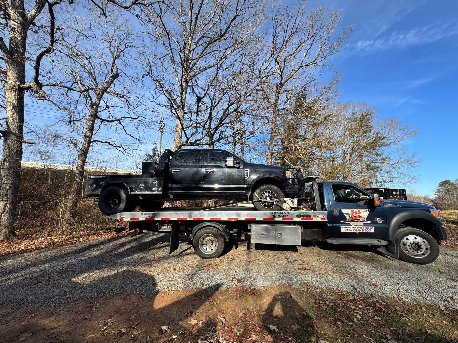 A black truck is on the flatbed of a tow truck in a wooded area on a sunny day.