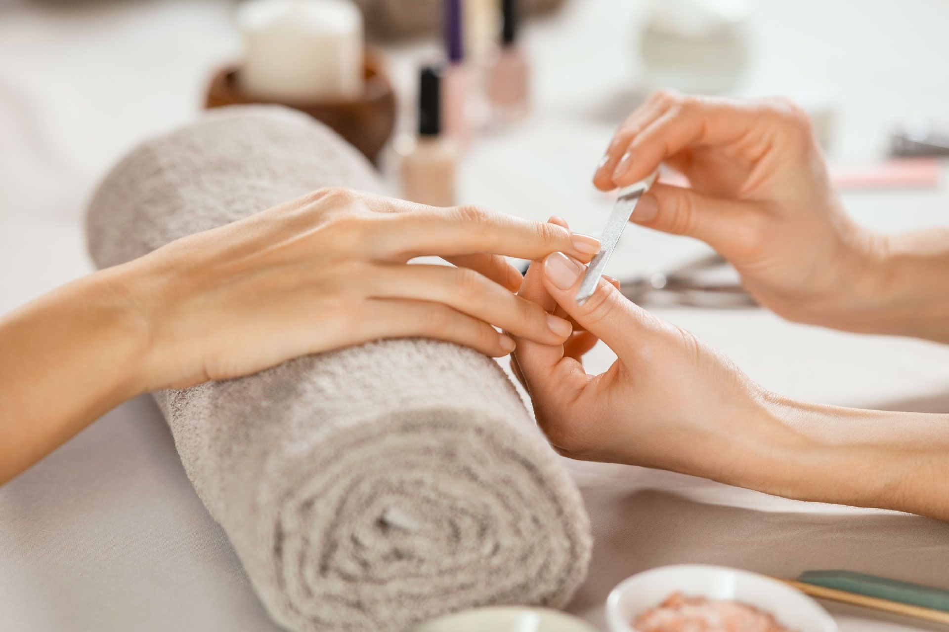 Manicurista pintando las uñas de una clienta en un spa. La mano de la clienta descansa sobre una toalla enrollada; hay botellas y herramientas cerca.