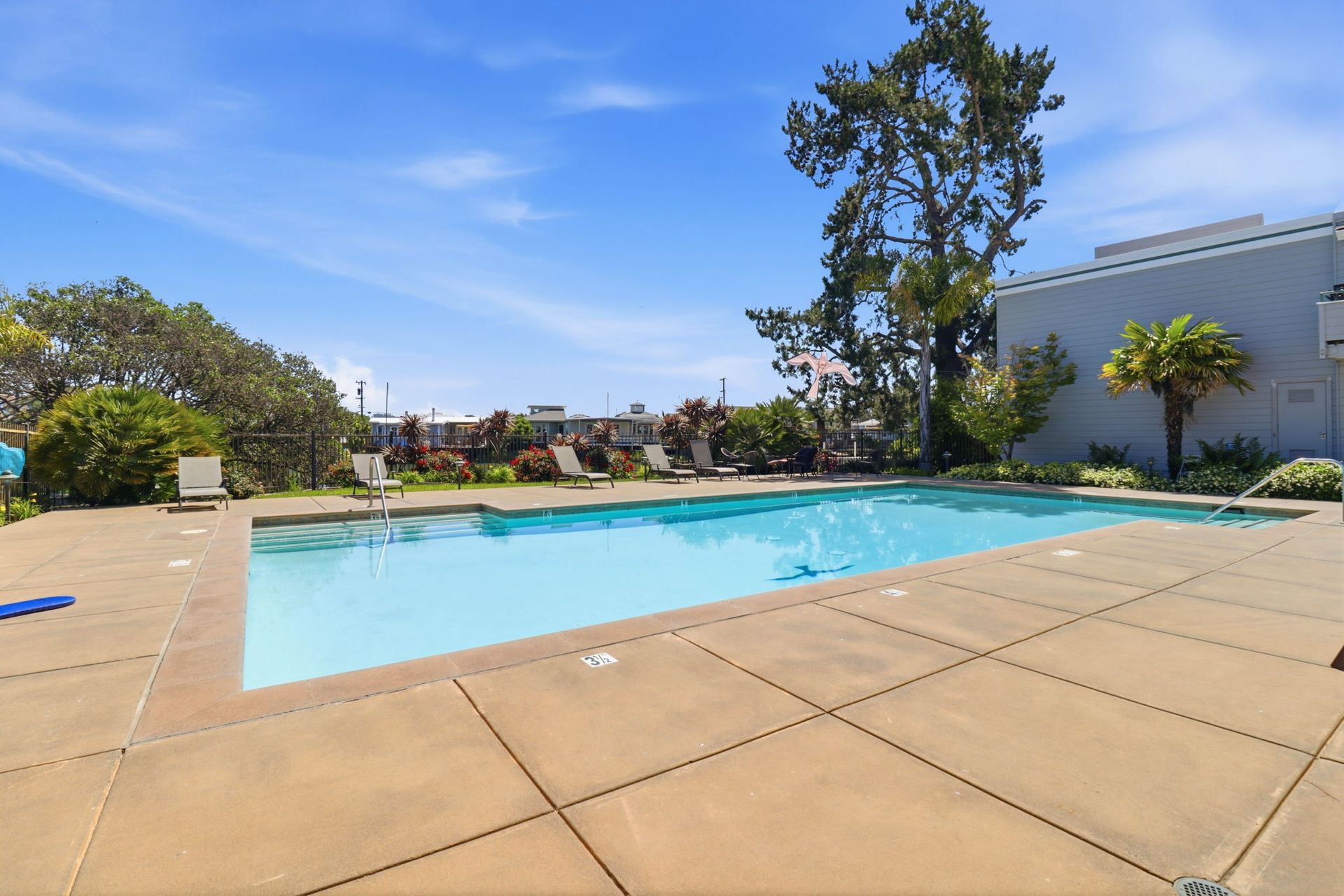 Outdoor swimming pool with tan deck, blue water, trees, and apartment buildings under a clear sky