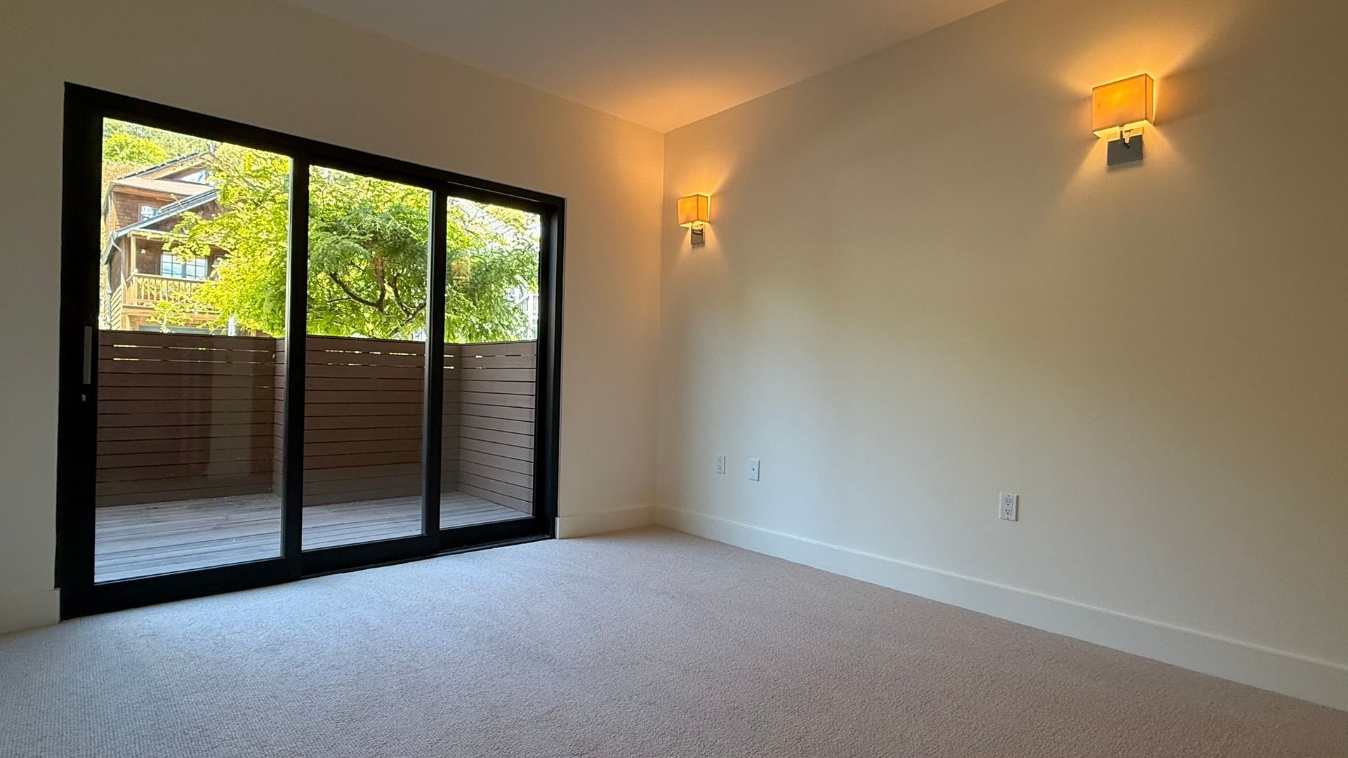 Empty bedroom with sliding glass door to a balcony, beige carpet, white walls, two sconces.