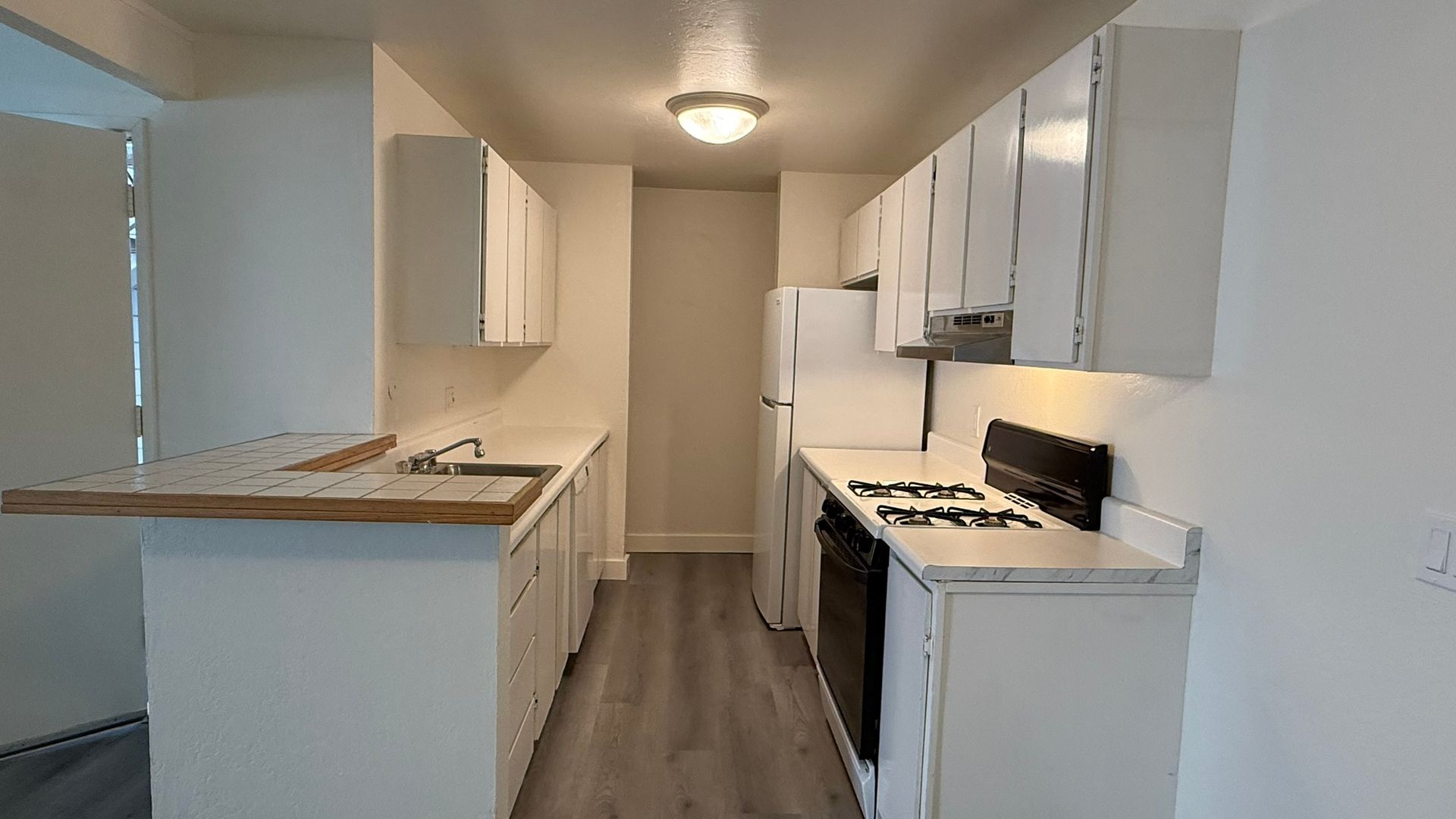 A small, white kitchen with appliances, cabinets, and a countertop, in a narrow apartment.