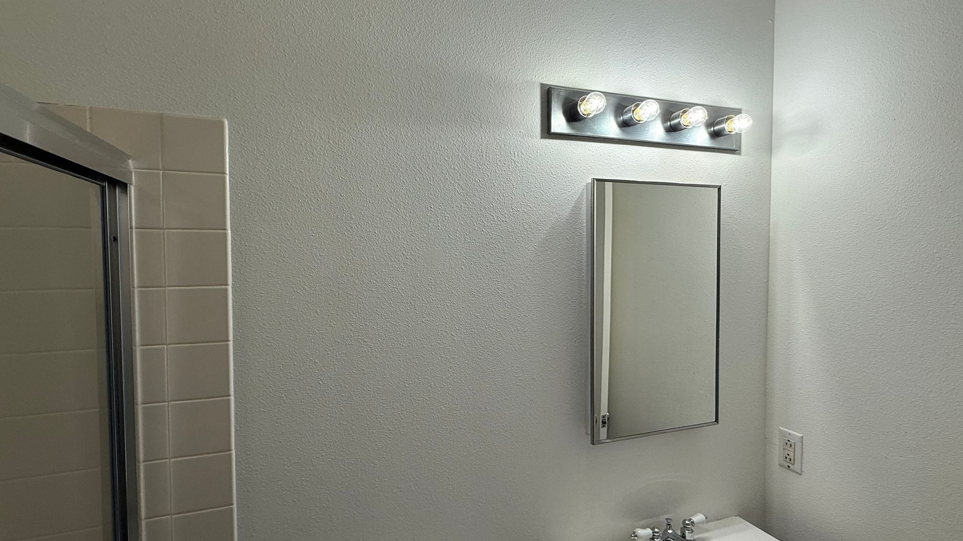 Bathroom with a mirror, light fixture, and shower stall; white walls, beige tiles.