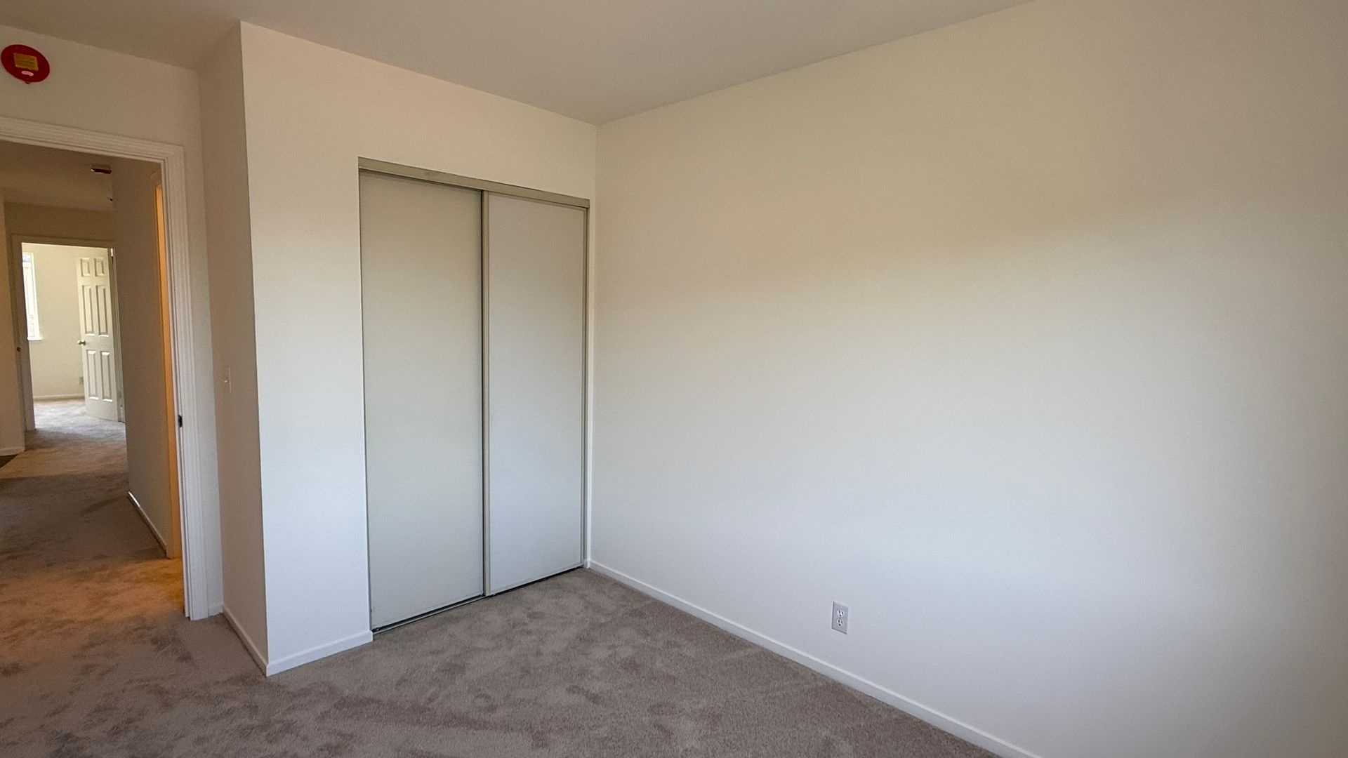 Empty bedroom with beige walls, closet, and carpet; an open doorway to the left.