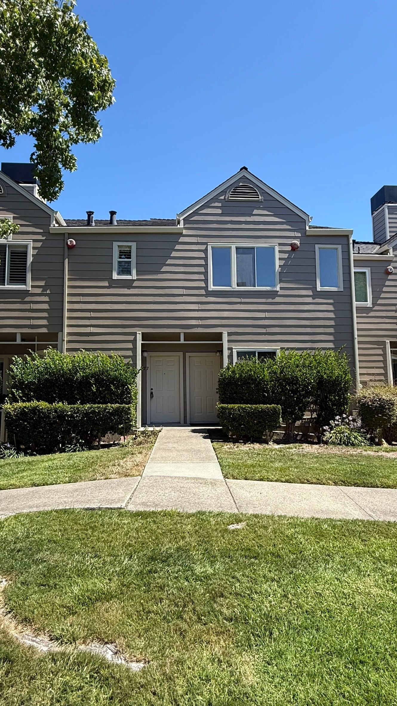 A gray, two-story townhouse with a white door and two small bushes.