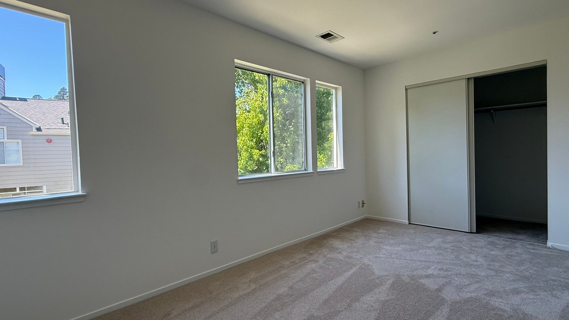 Empty room with tan carpet, three windows, and a closet; light gray walls.