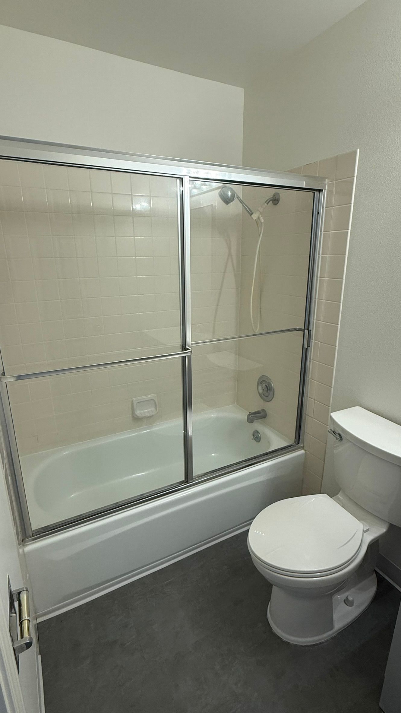 Bathroom with white bathtub, sliding glass shower door, toilet, and gray flooring.