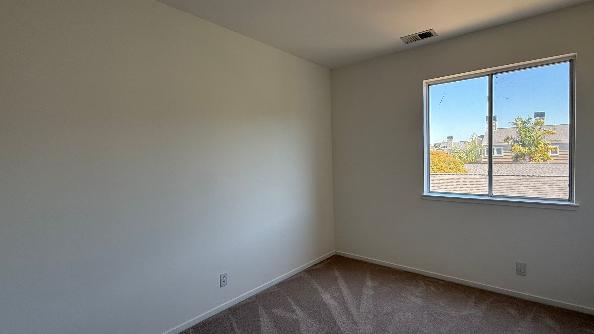 Empty room with tan carpet, off-white walls, and a window with a view of a bright, sunny day.