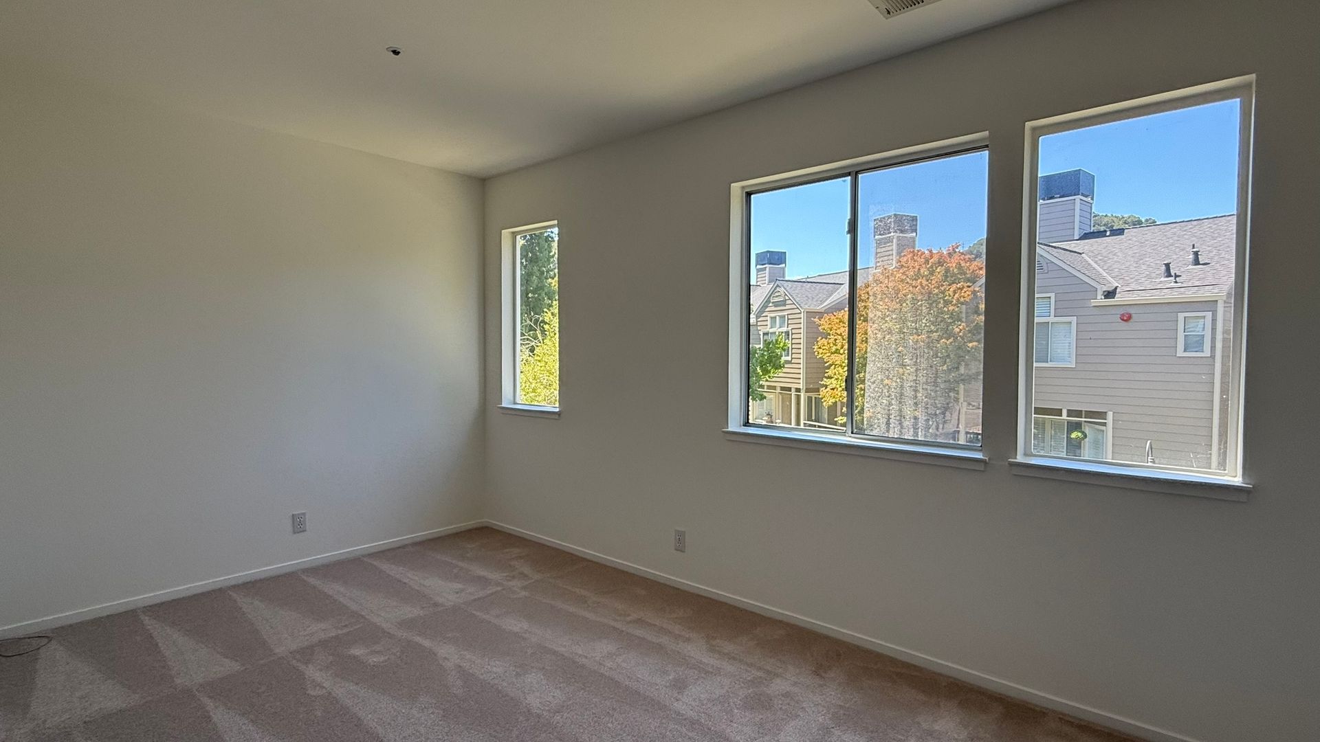 Empty room with beige carpet and three windows overlooking a building.
