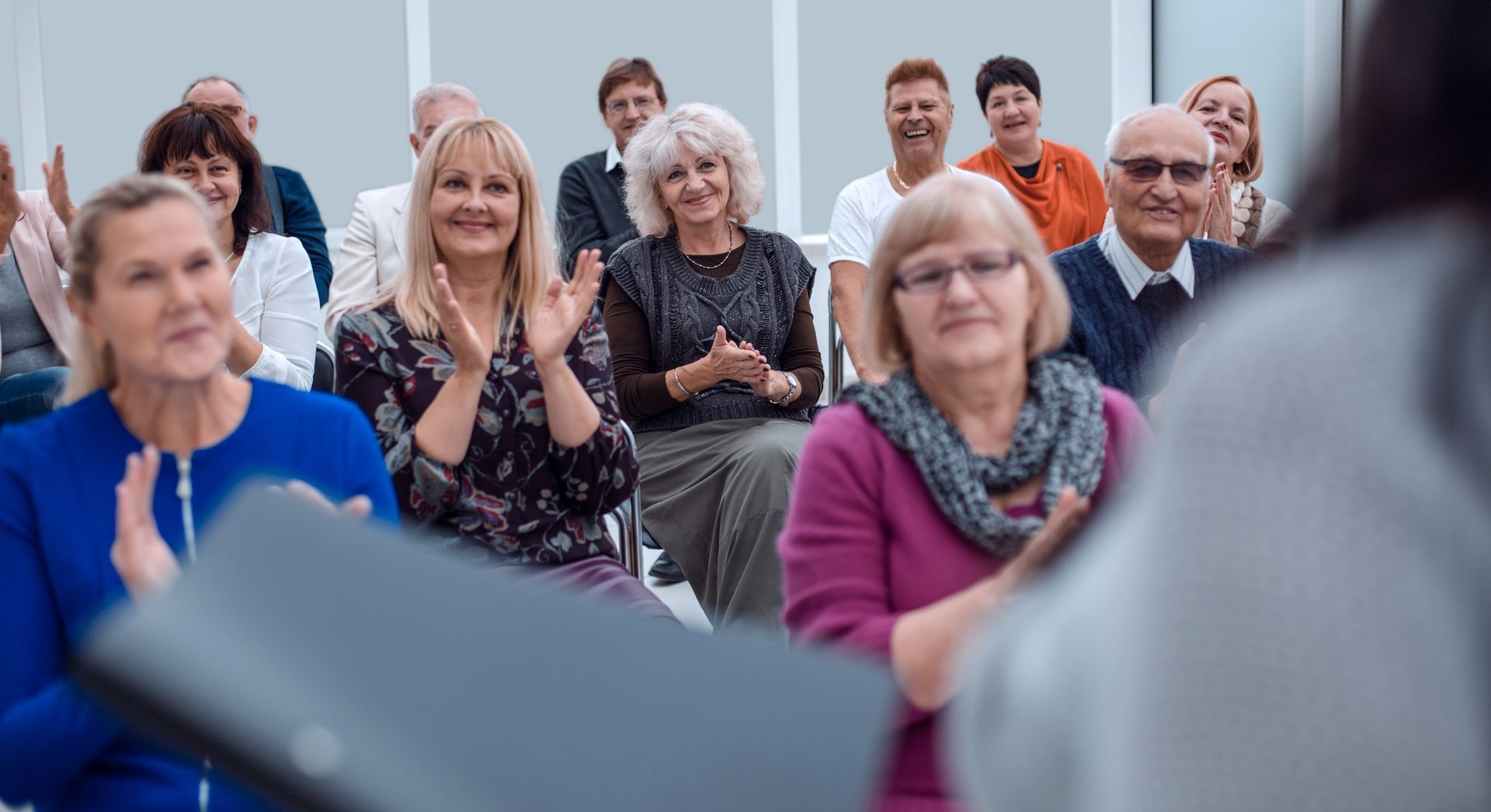 Audience applauds speaker at a presentation. Many are smiling. Indoors, seated audience.