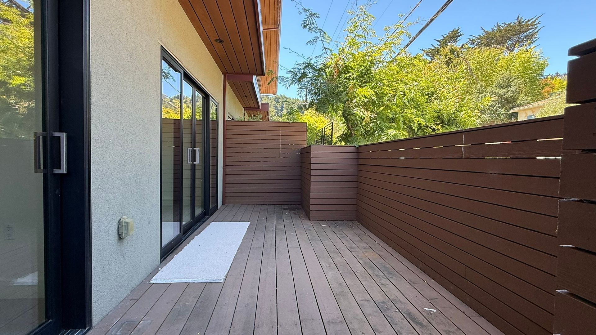 Wooden deck with a brown slatted fence, next to a white wall and sliding glass doors.