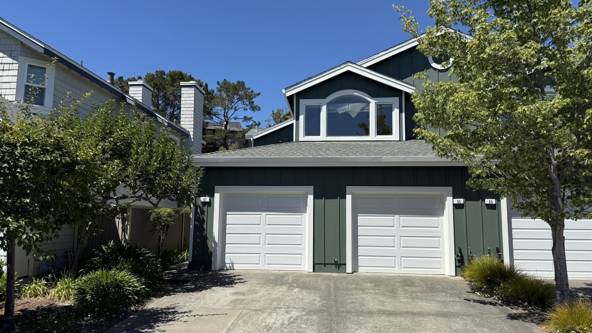 Green house with white garage doors, driveway, and trees under a blue sky.