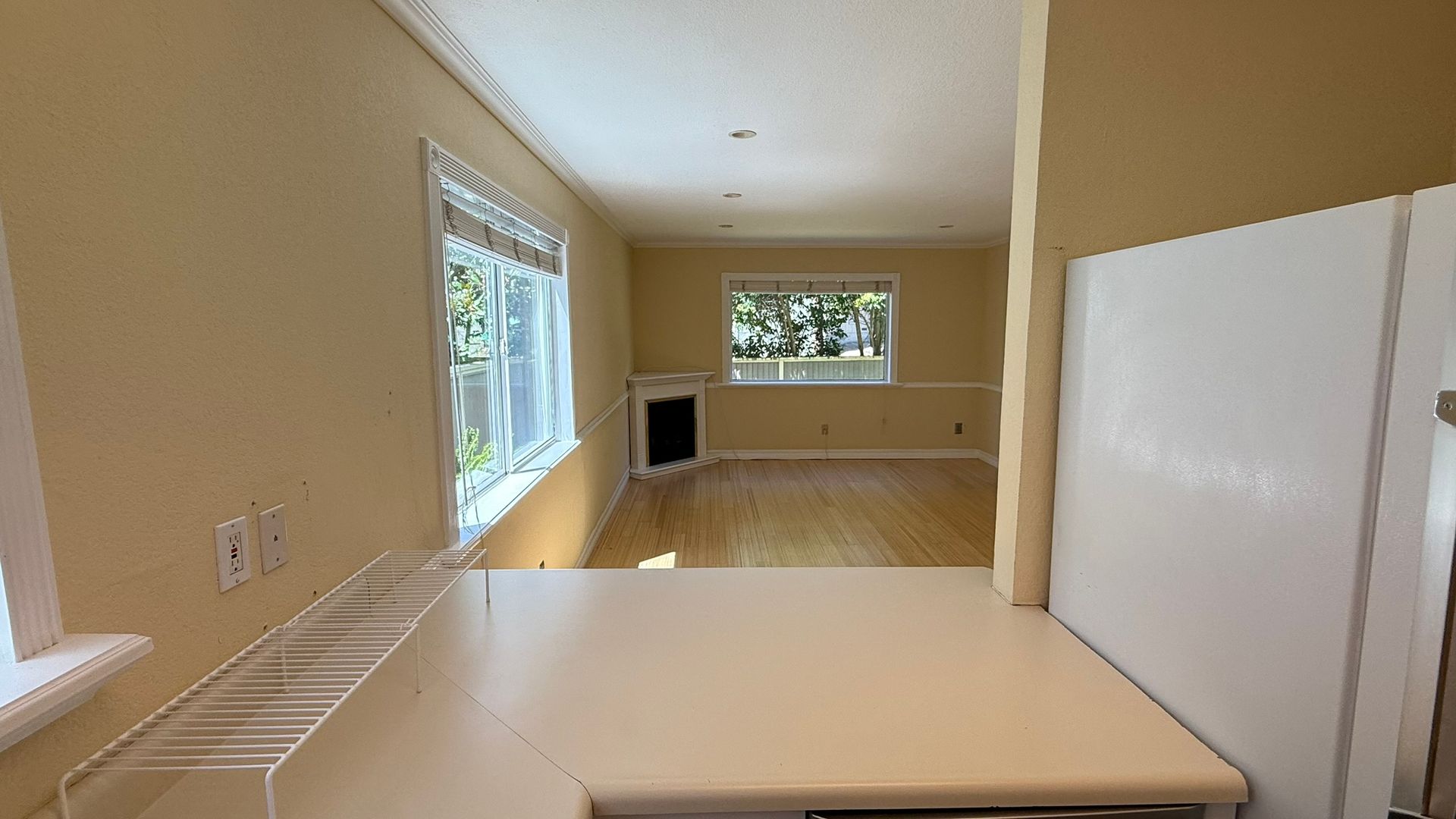 View from kitchen counter into a living room with windows, light walls, and wood floors.