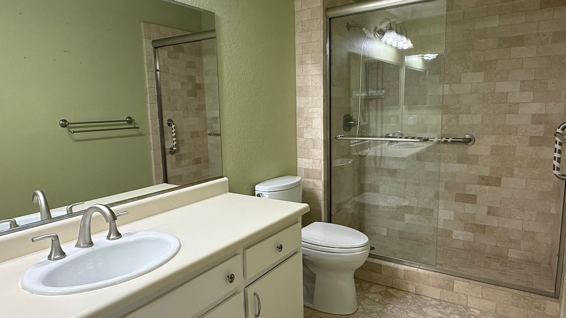 Bathroom with a white vanity, toilet, and a shower with grab bars. The walls are pale green and beige.