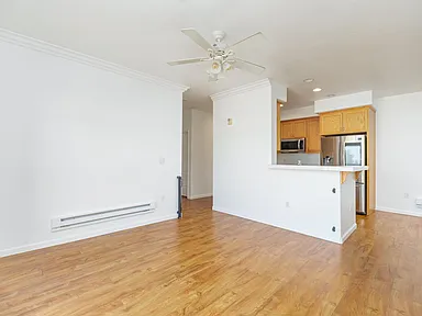 Empty living space with wood floors, white walls, partial kitchen, and a ceiling fan.