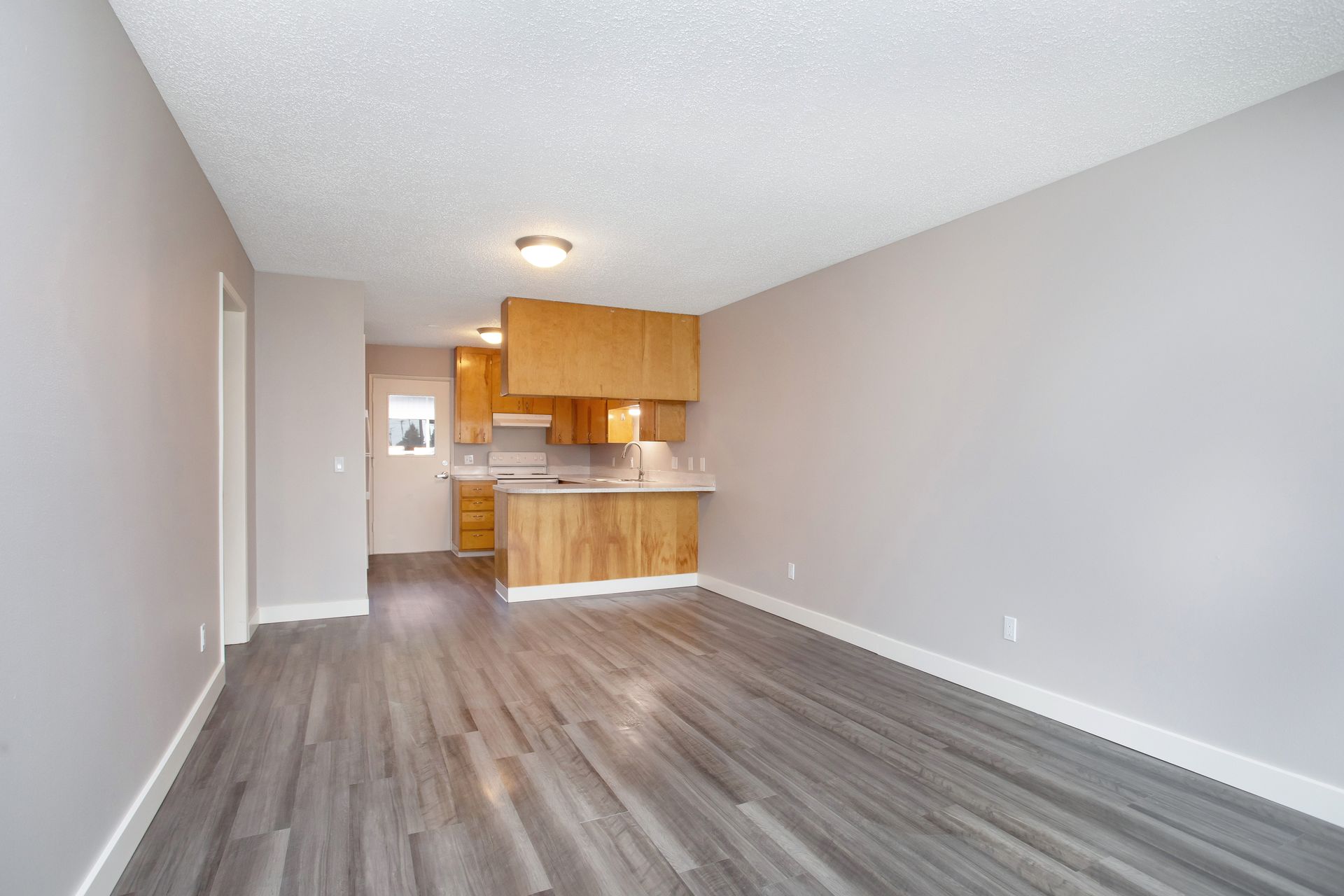 An empty living room with hardwood floors and a kitchen in the background.