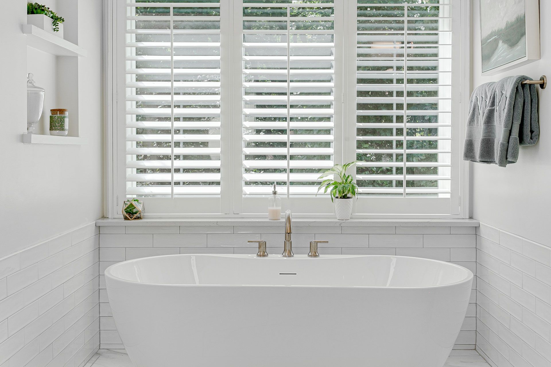 White bathroom with a freestanding tub under a window with white shutters.