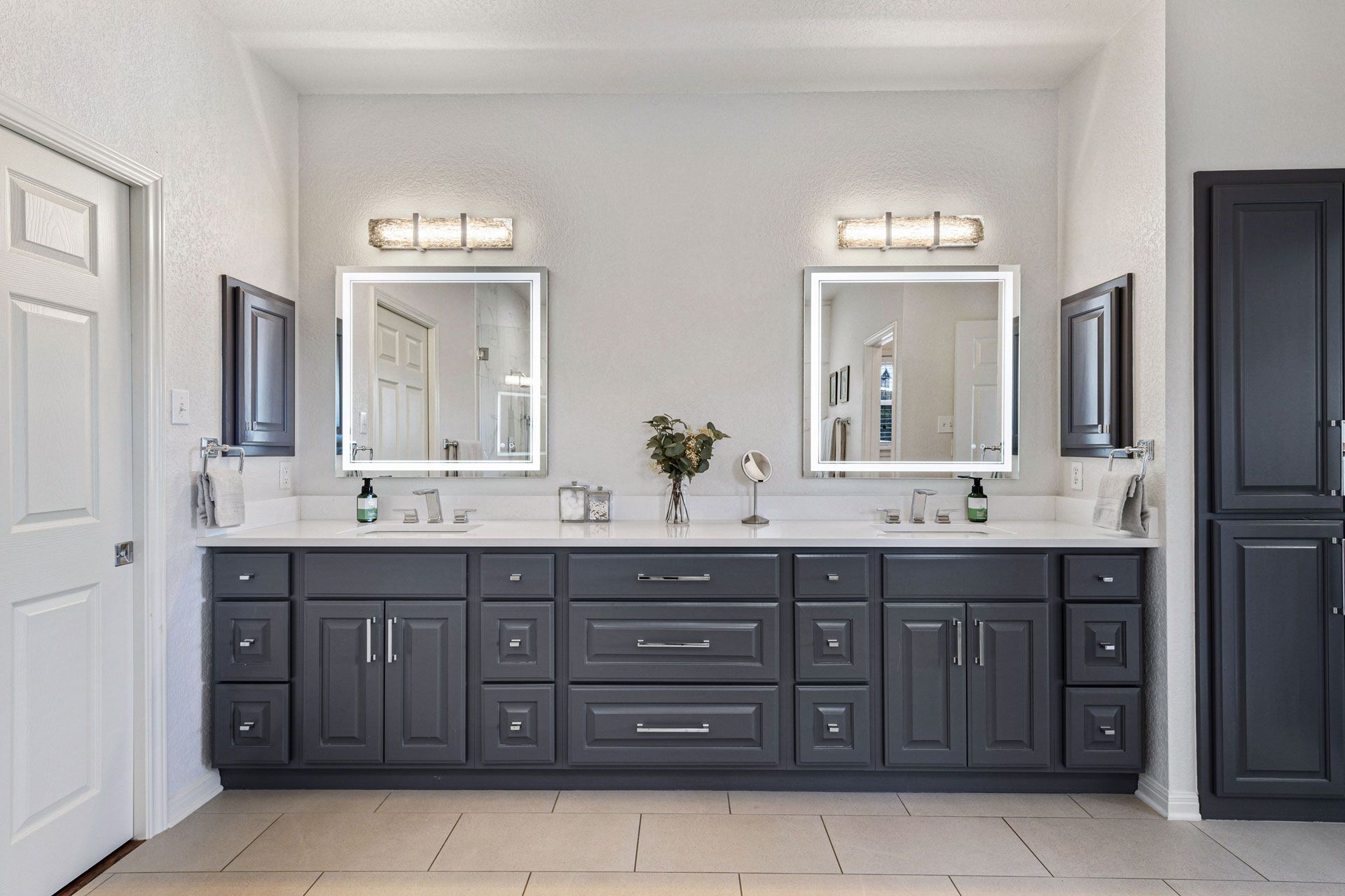 Bathroom with two sinks, dark gray cabinets, white countertops, and illuminated mirrors.
