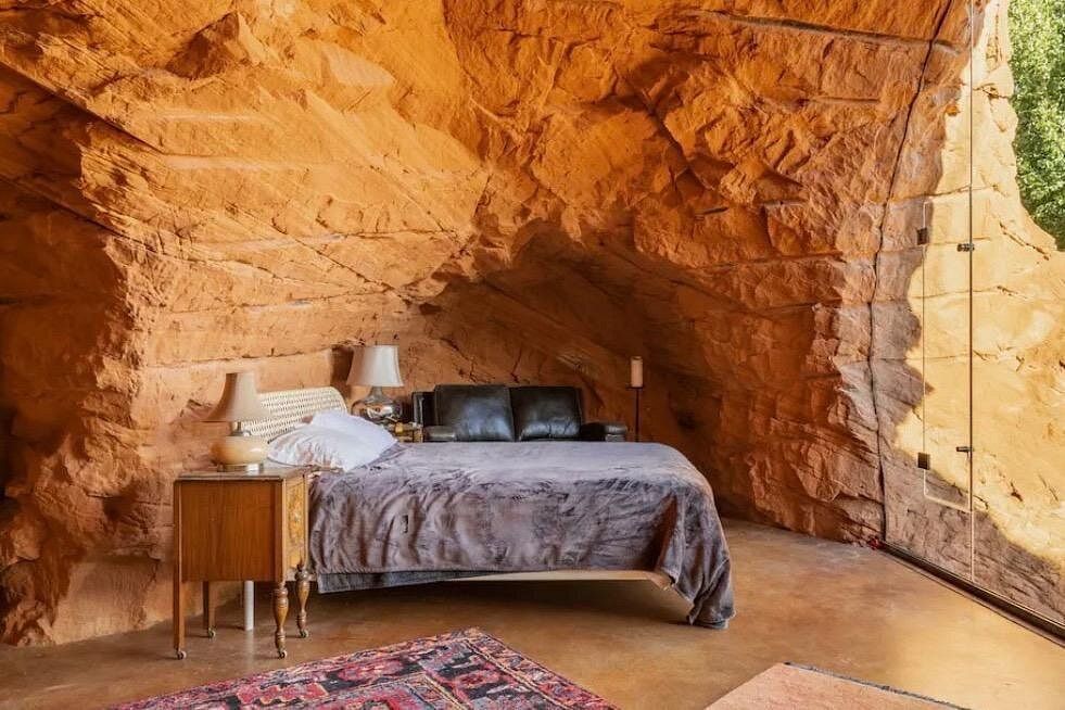Bedroom built into red rock, bed with gray blanket, wood nightstand, rug, and large window.