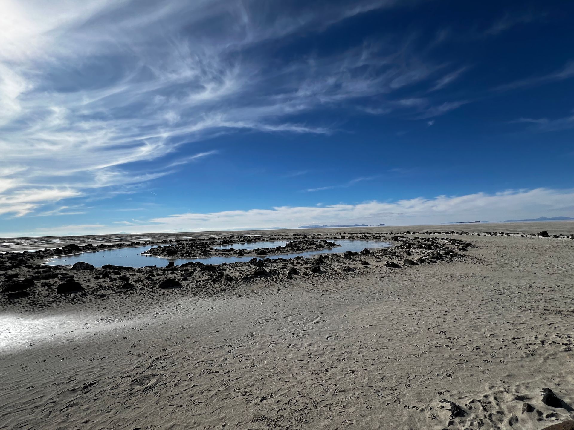 Barren, light-colored landscape under a bright blue sky with wispy clouds. A dark, rocky formation holds shallow water.