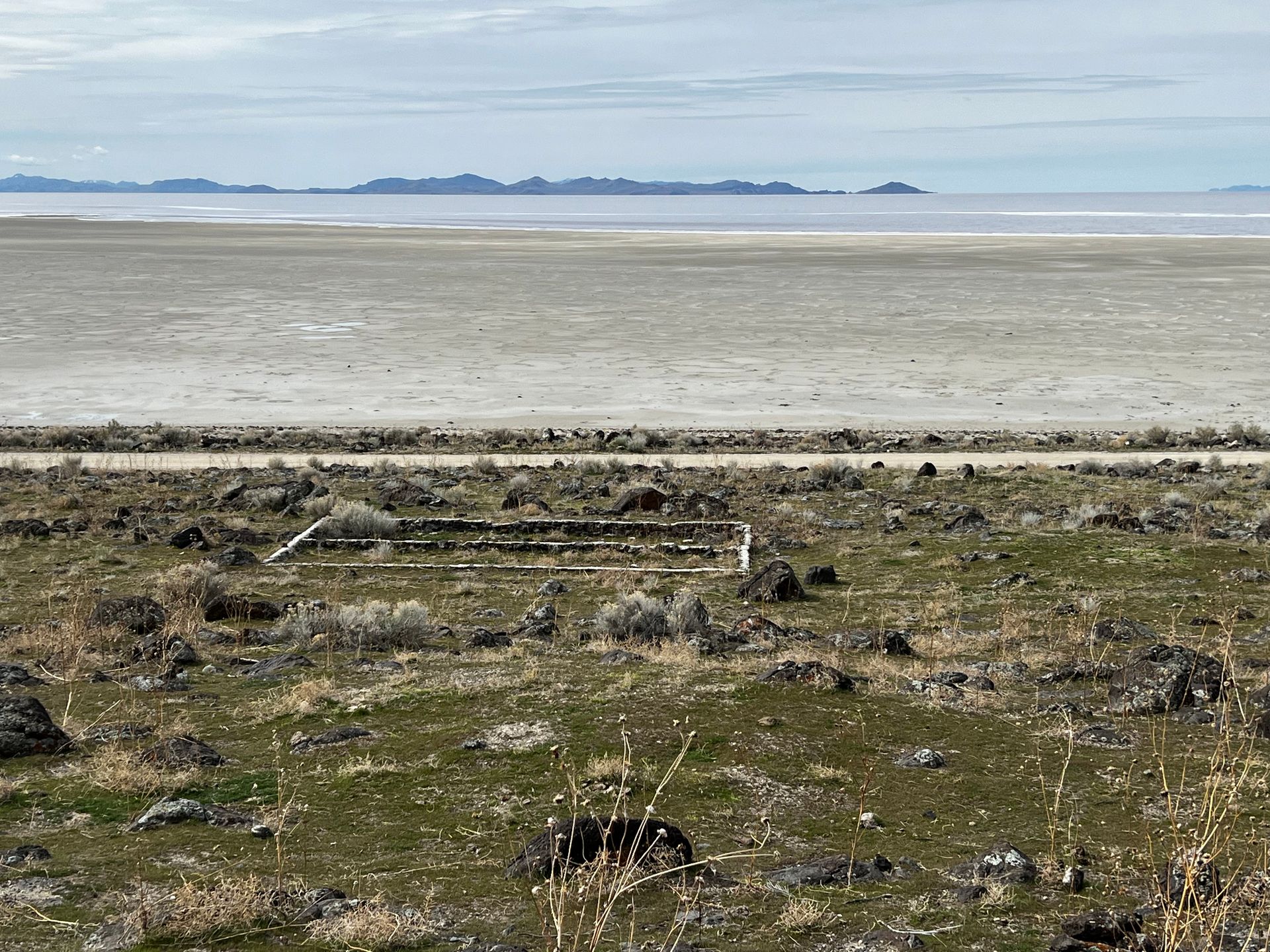 Barren landscape with salt flats, a small concrete structure, and distant mountains under a cloudy sky.