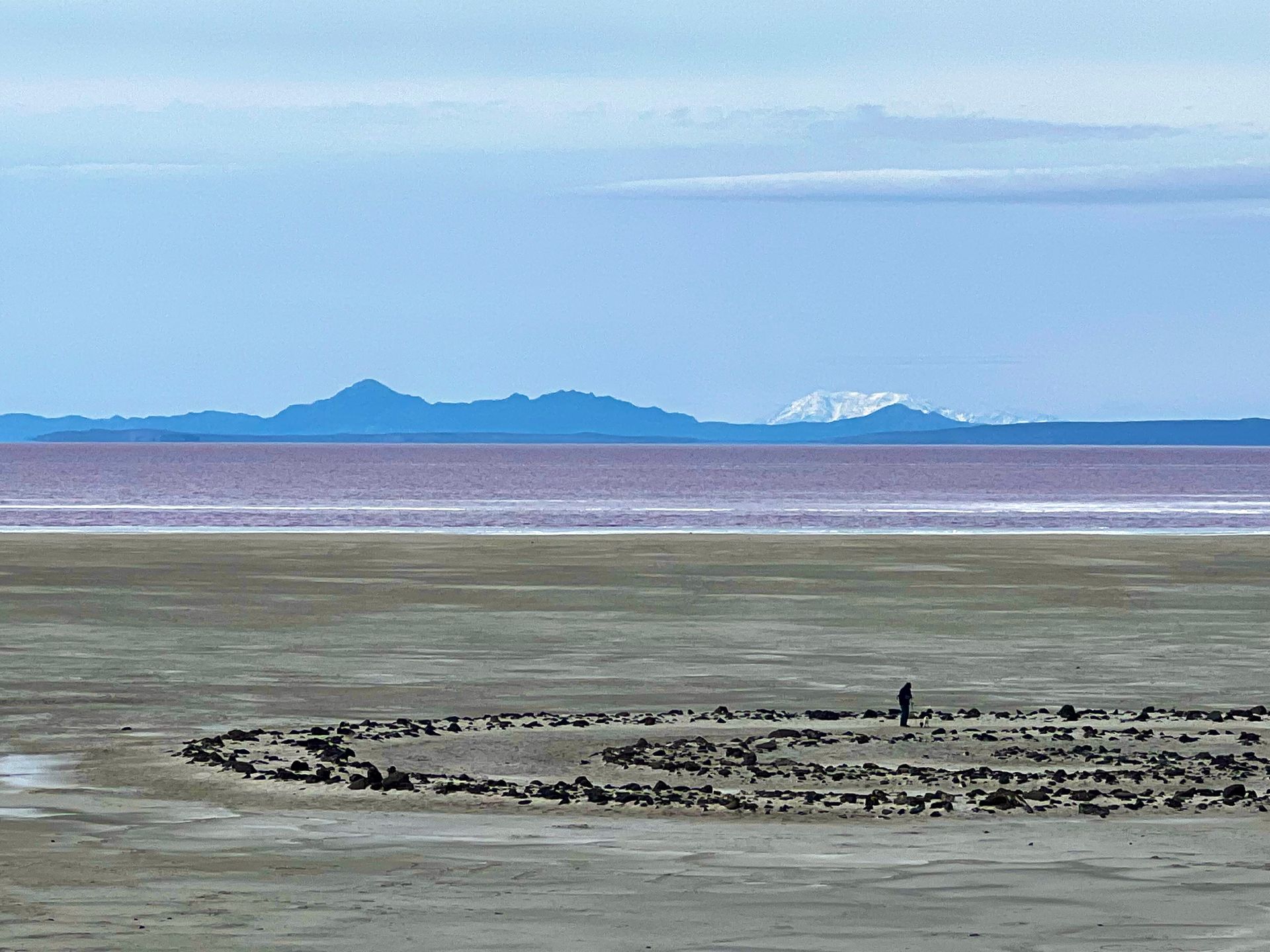Person stands in a rock circle on a grey plain with pink water and mountains under a blue sky.