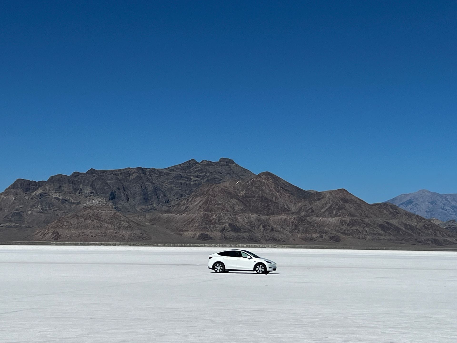 White car on a vast, white salt flat, with a mountain range in the background under a blue sky.