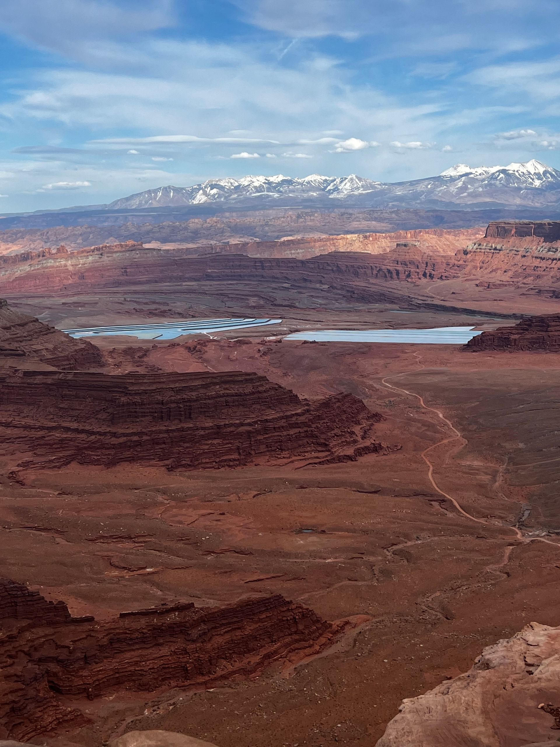 Red desert landscape with a blue-tinged lake, mountains in the distance, and a partly cloudy sky.