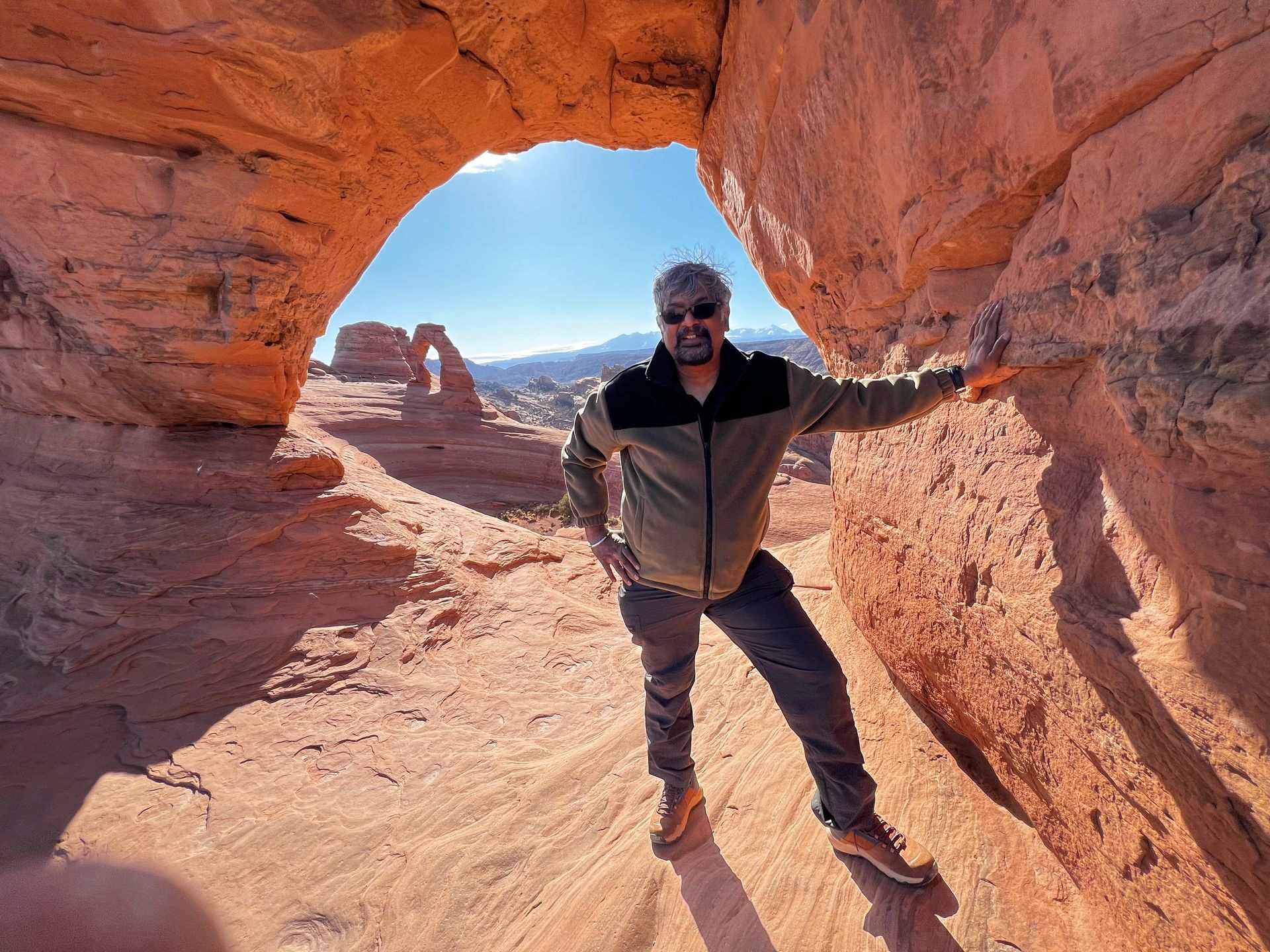 Man standing inside a red rock arch, sunshine, brown jacket, blue sky, desert landscape.