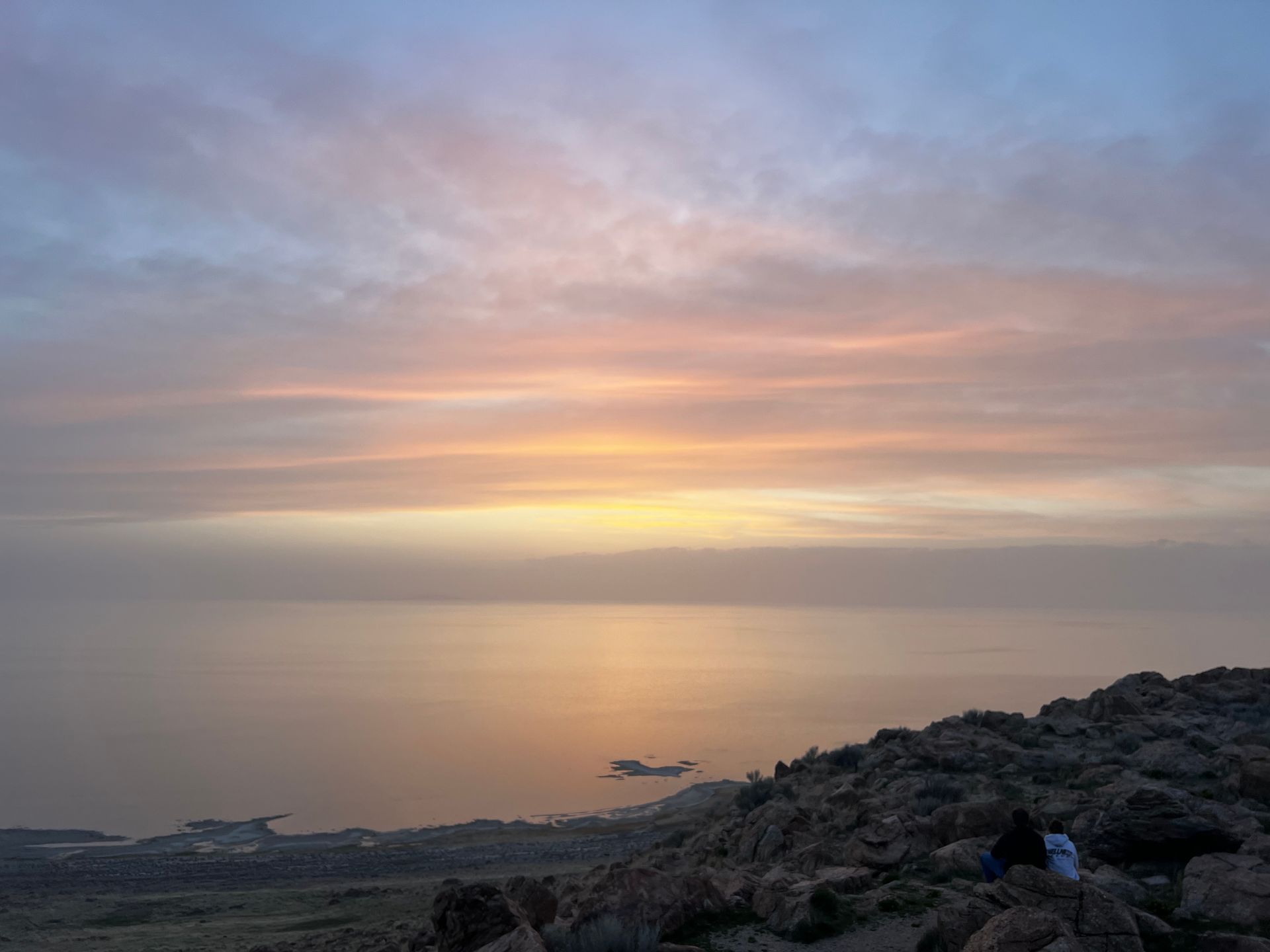 Sunset over a calm body of water, viewed from a rocky mountaintop; soft oranges and pinks in the sky.