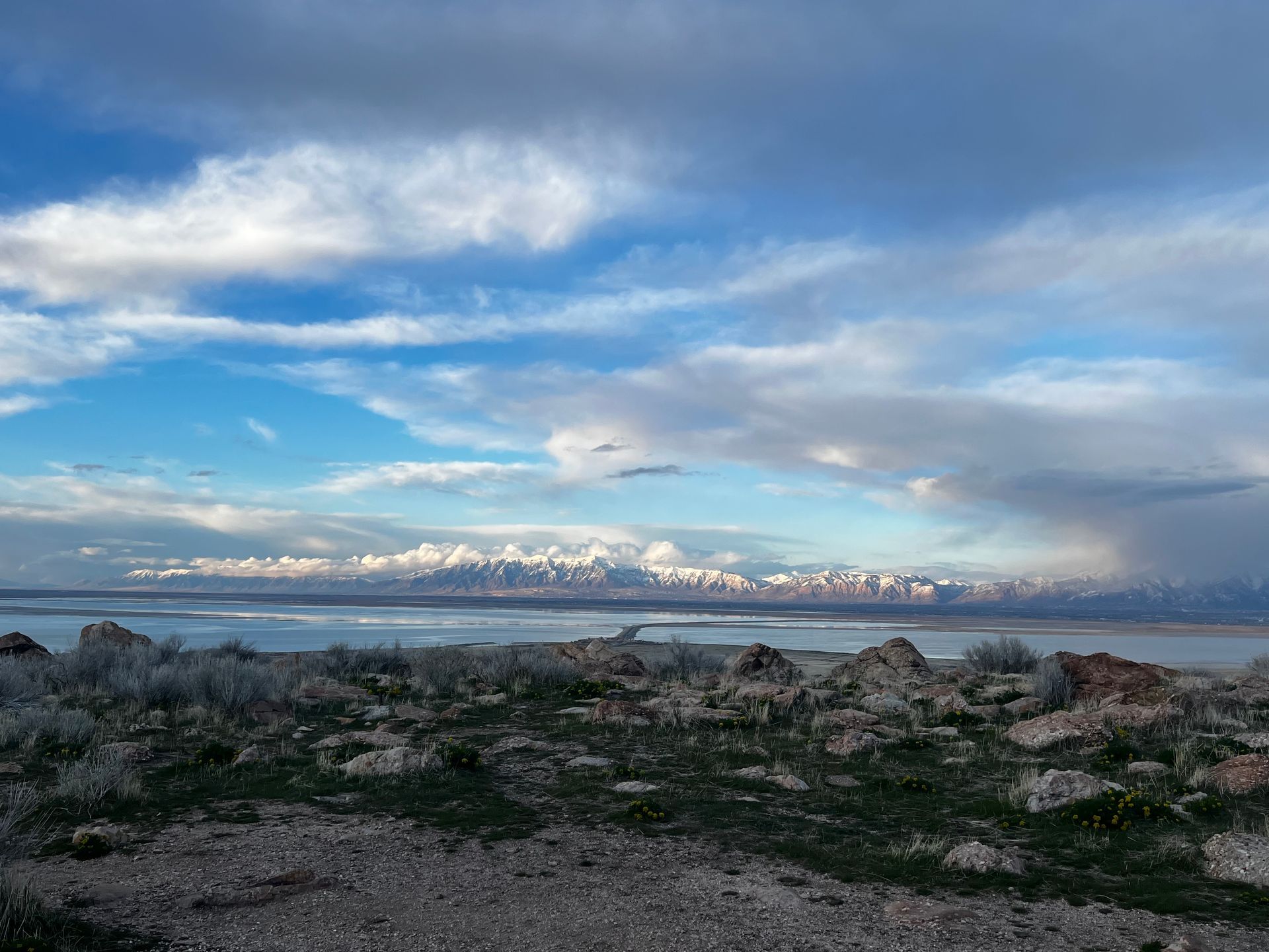 Overlooking a lake, a rocky foreground leads to distant snow-capped mountains under a cloudy blue sky.