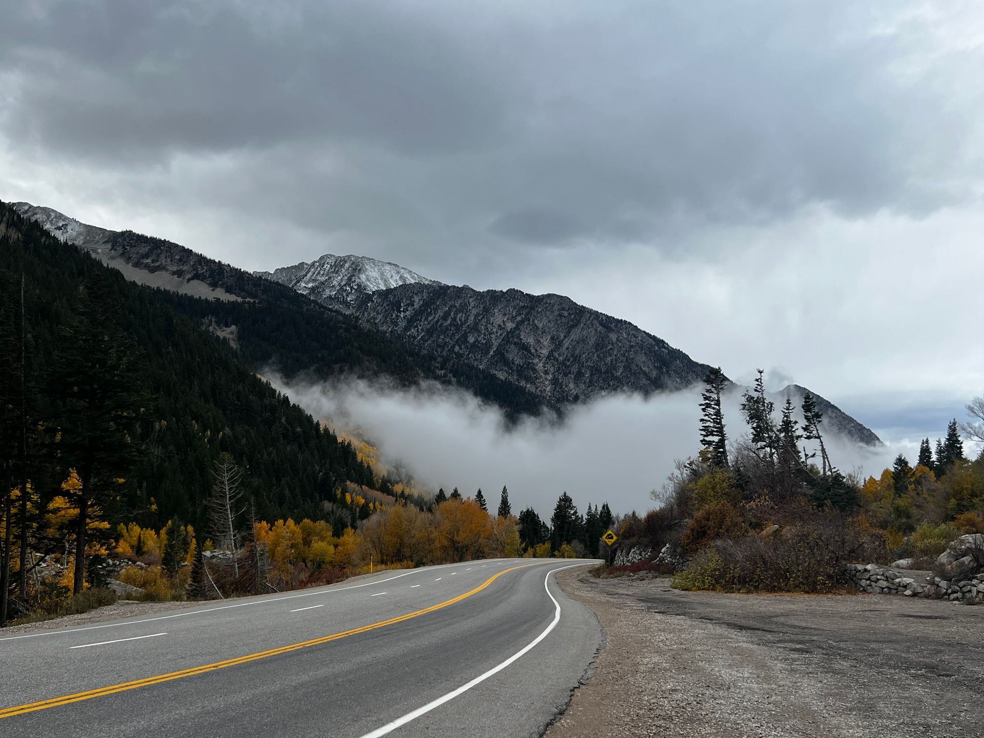 Road winds through mountain landscape with cloudy sky. Fall colors along the road and snow-capped peaks.