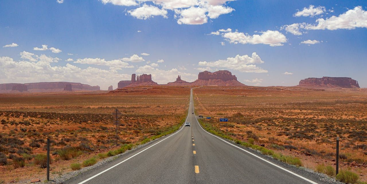 A long, straight road through a desert landscape, with red rock formations in the distance under a blue sky.