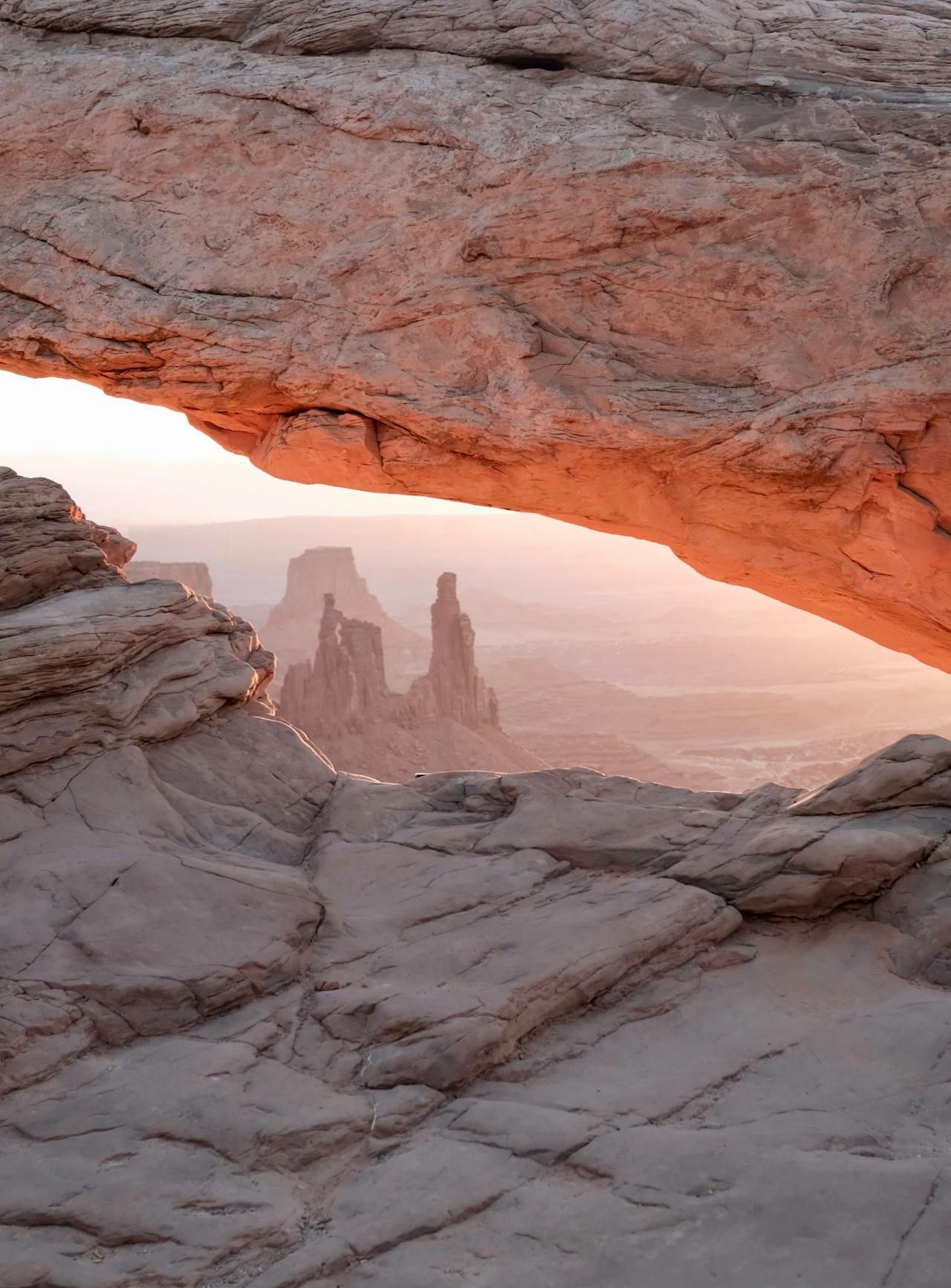 Sandstone arch frames a distant, hazy landscape with red-orange sunset.