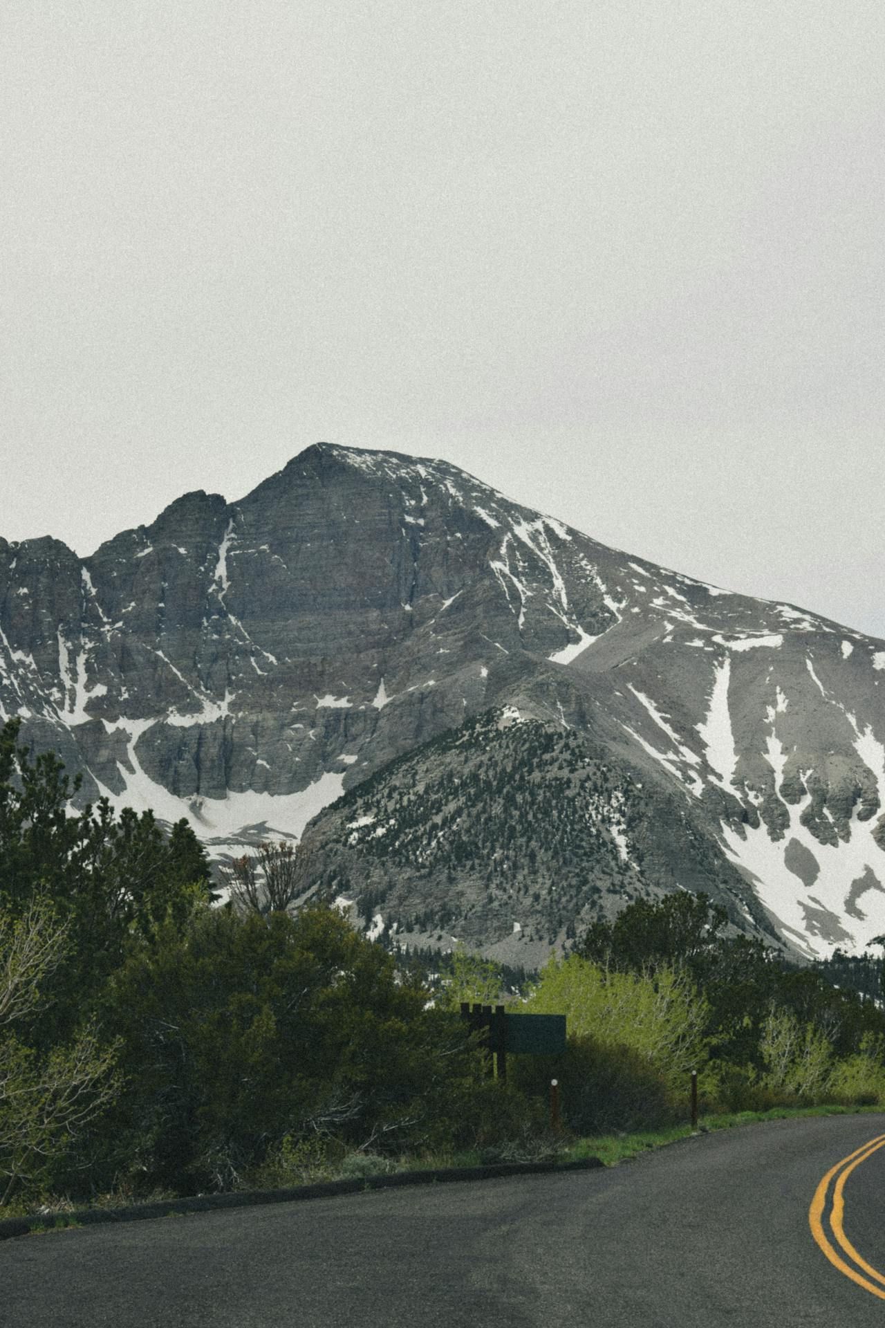 Snow-capped mountain rising above a winding road with trees, against a cloudy sky.